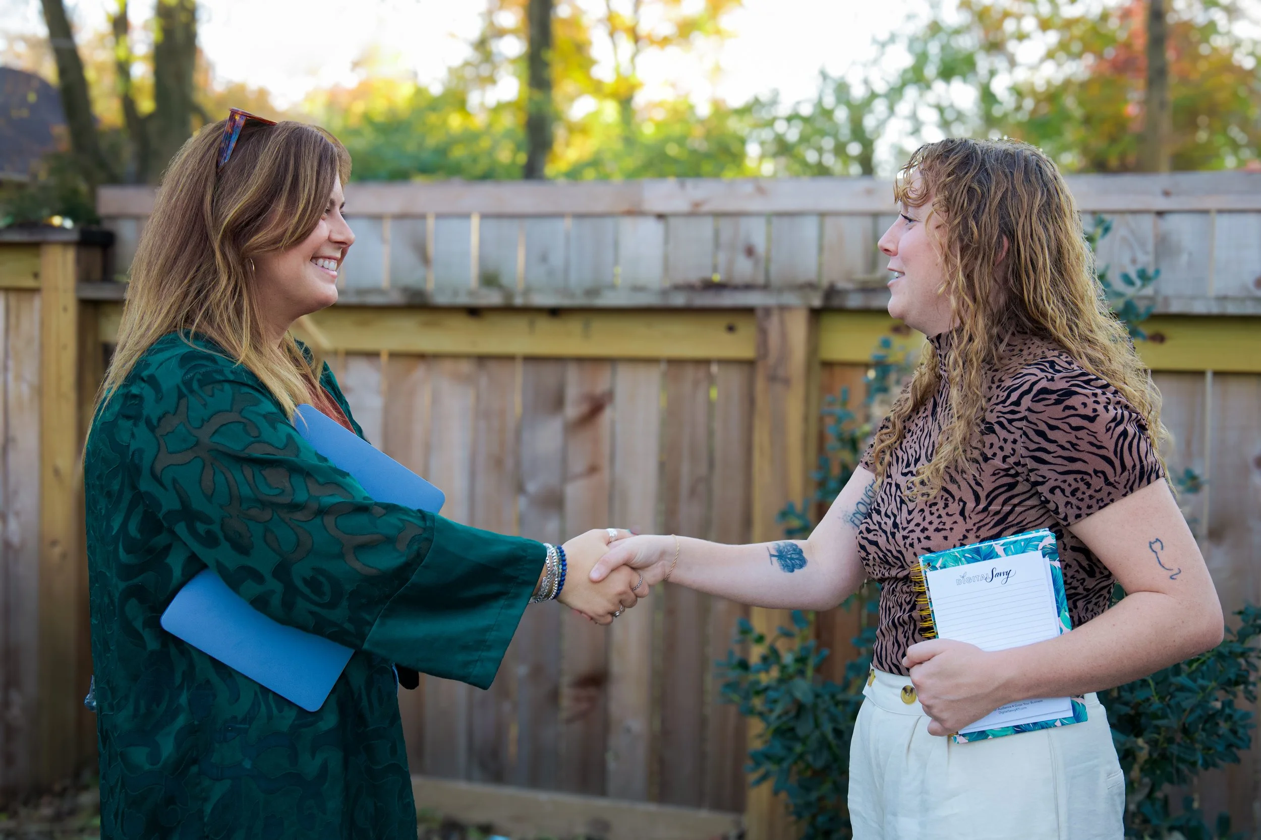 Two women shaking hands outdoors, smiling, with a wooden fence and trees in the background.