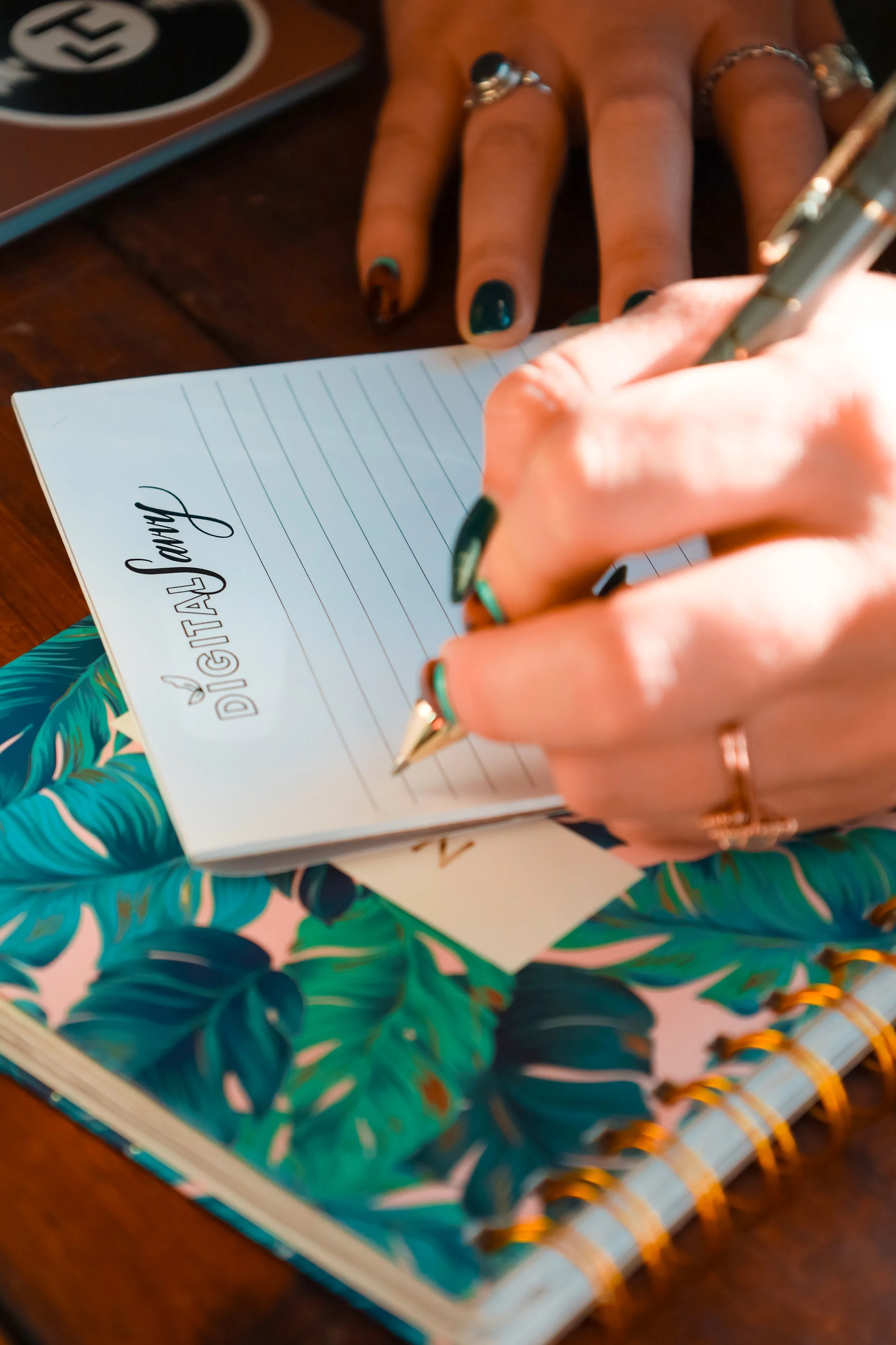 A person is writing a note on a notepad titled 'Digital Journal' with a pen. The person has colorful painted nails and is wearing rings and a bracelet. The notepad has a tropical leaf design on the cover.