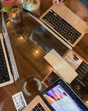 A wooden table with a laptop, tablet, a glass of iced tea with a lemon slice, a menu, and some papers. There are two people in the scene, one with light-colored nails and another with a hand on the laptop.