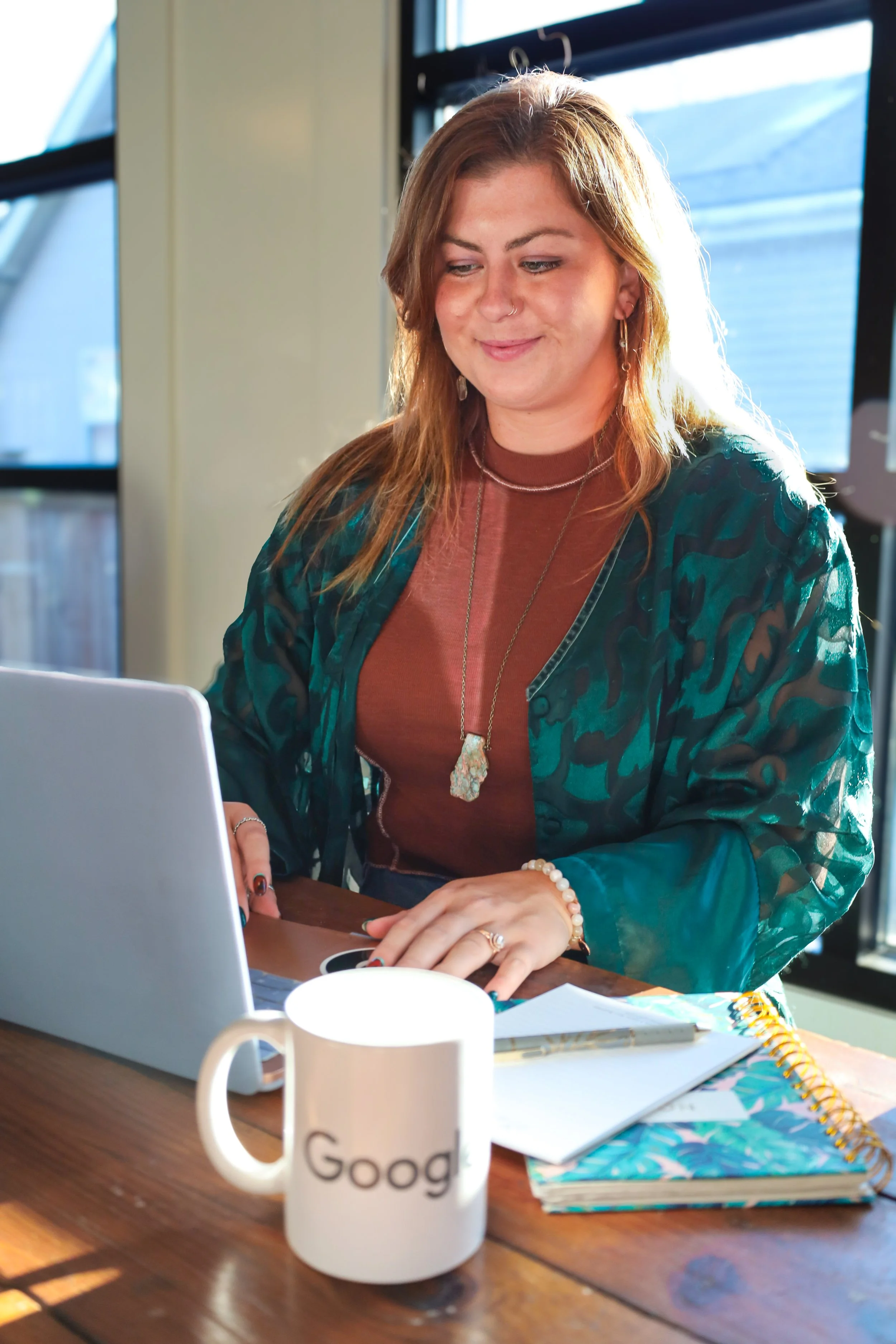 A woman sitting at a wooden table in front of a laptop, with a white mug, notebooks, and a pen, working in a bright room with large windows.