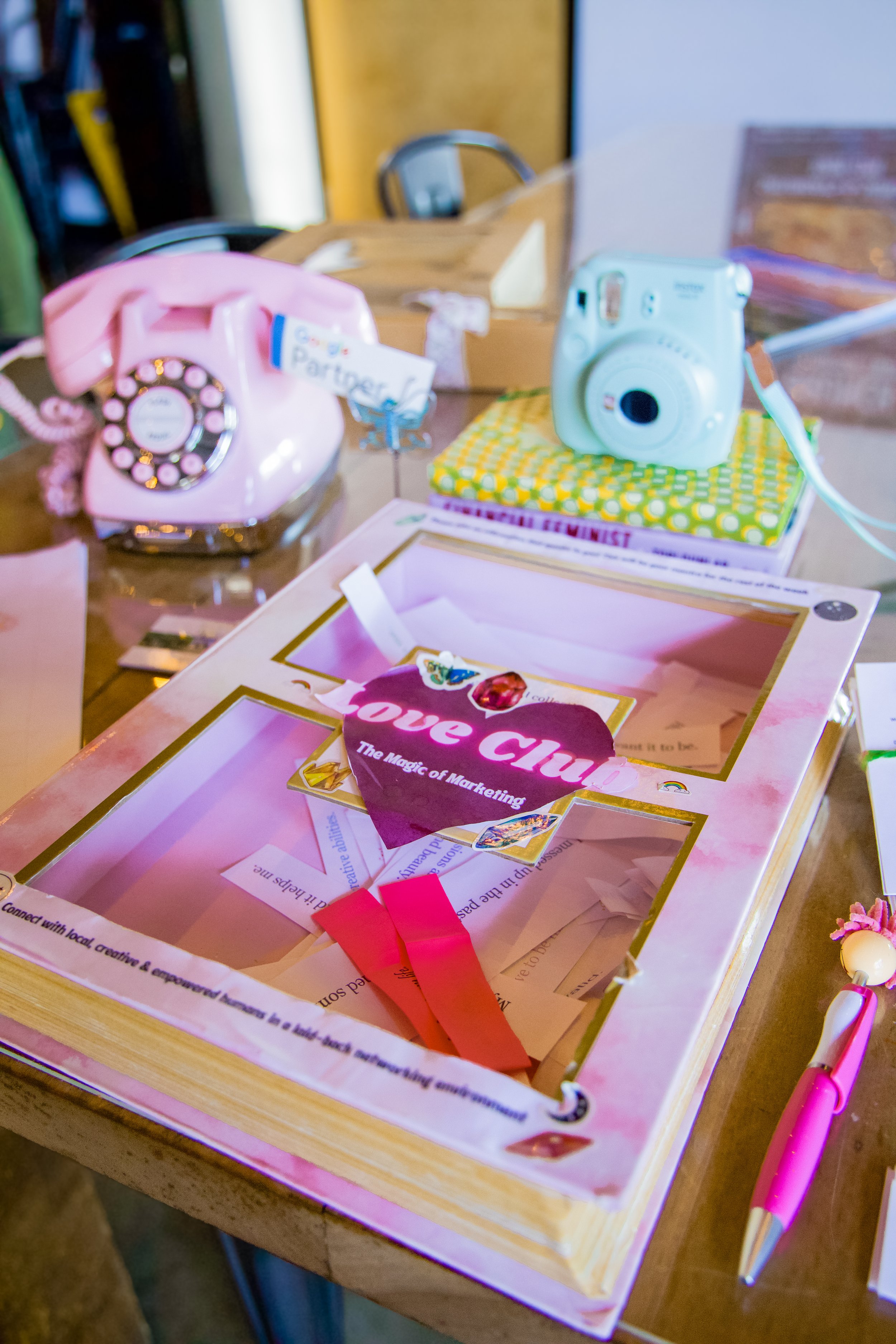 A cluttered desk with a pink rotary telephone, a mint-green instant camera, a pink pen, and a pink display box with the words 'Love Club' and 'The Magic of Marketing' inside.