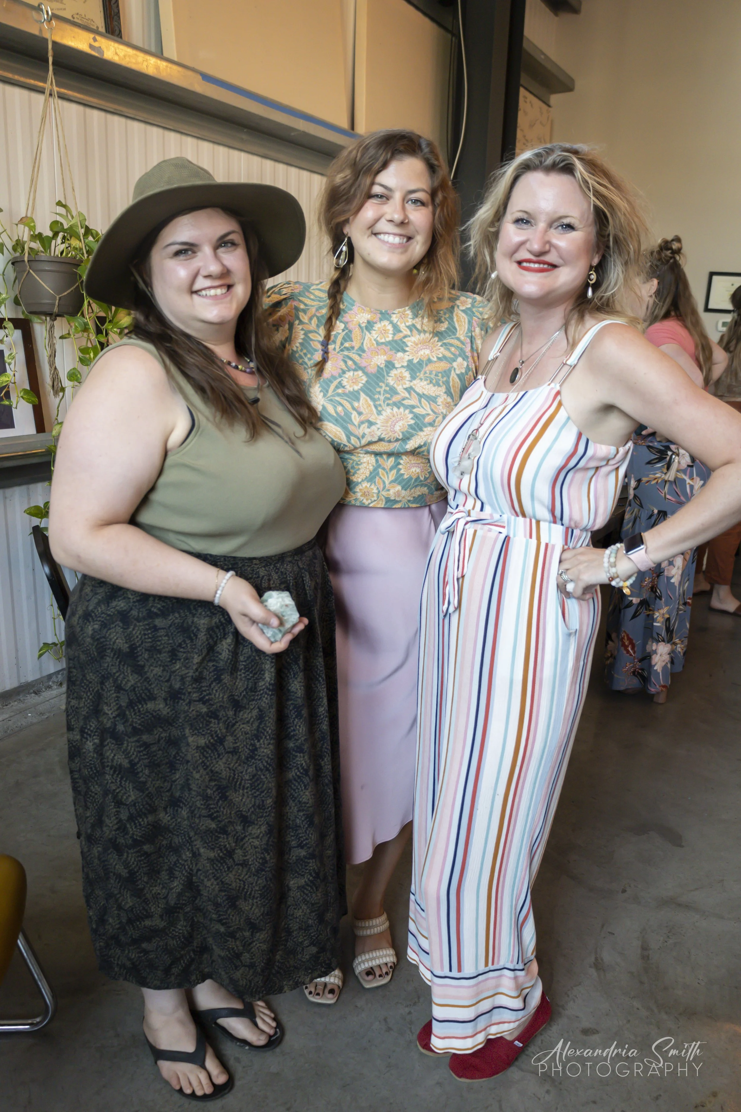 Three women standing together at an indoor gathering, smiling at the camera. The woman on the left is wearing a large tan hat, olive sleeveless top, long black patterned skirt, and sandals. The woman in the middle has wavy hair, a floral top, and pink skirt. The woman on the right has blonde curly hair, striped jumpsuit, and red shoes, with her hand on her hip.