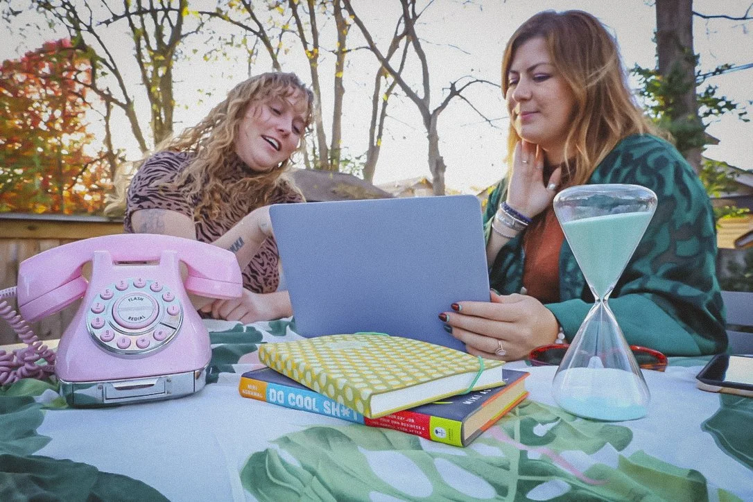 Two women are sitting at an outdoor table, looking at a laptop. One woman is smiling and the other is touching her neck. The table has a pink rotary phone, a yellow notebook, two books, an hourglass, and a smartphone. Background shows trees and a fence.