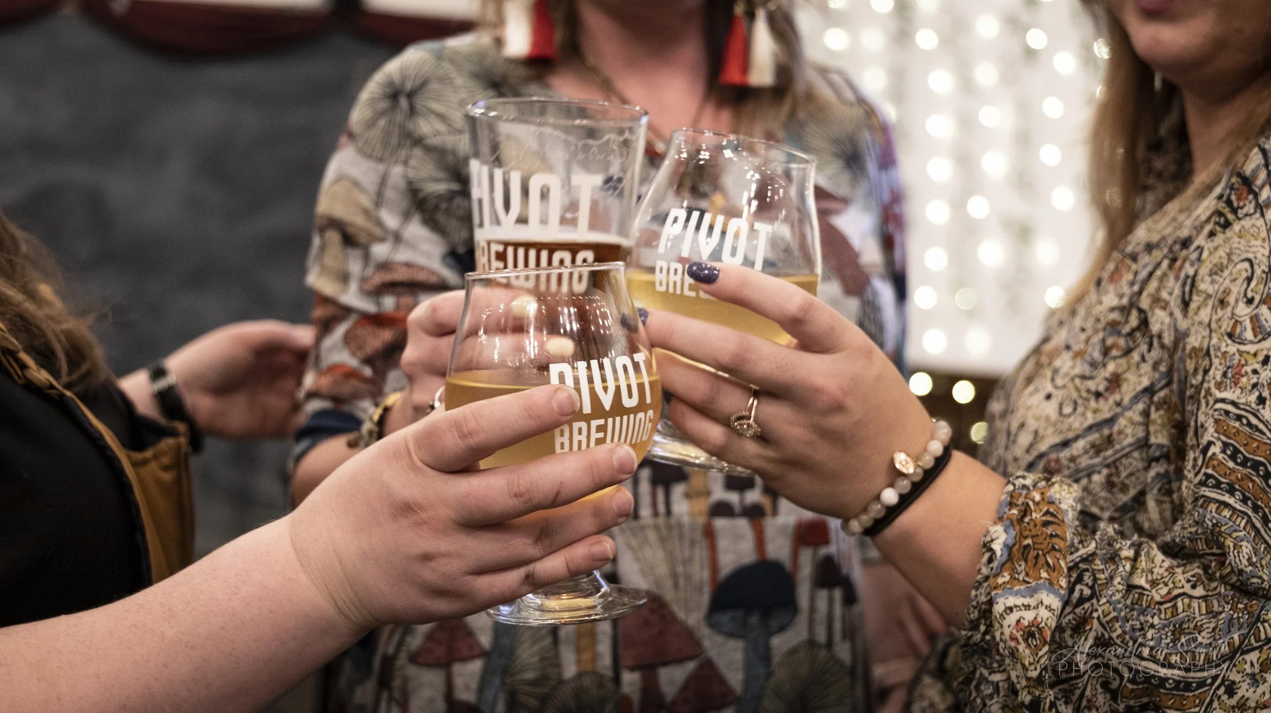 People clinking glasses of beer, with the logo 'Pivot Brewing' on the glasses, in a festive setting with string lights in the background.