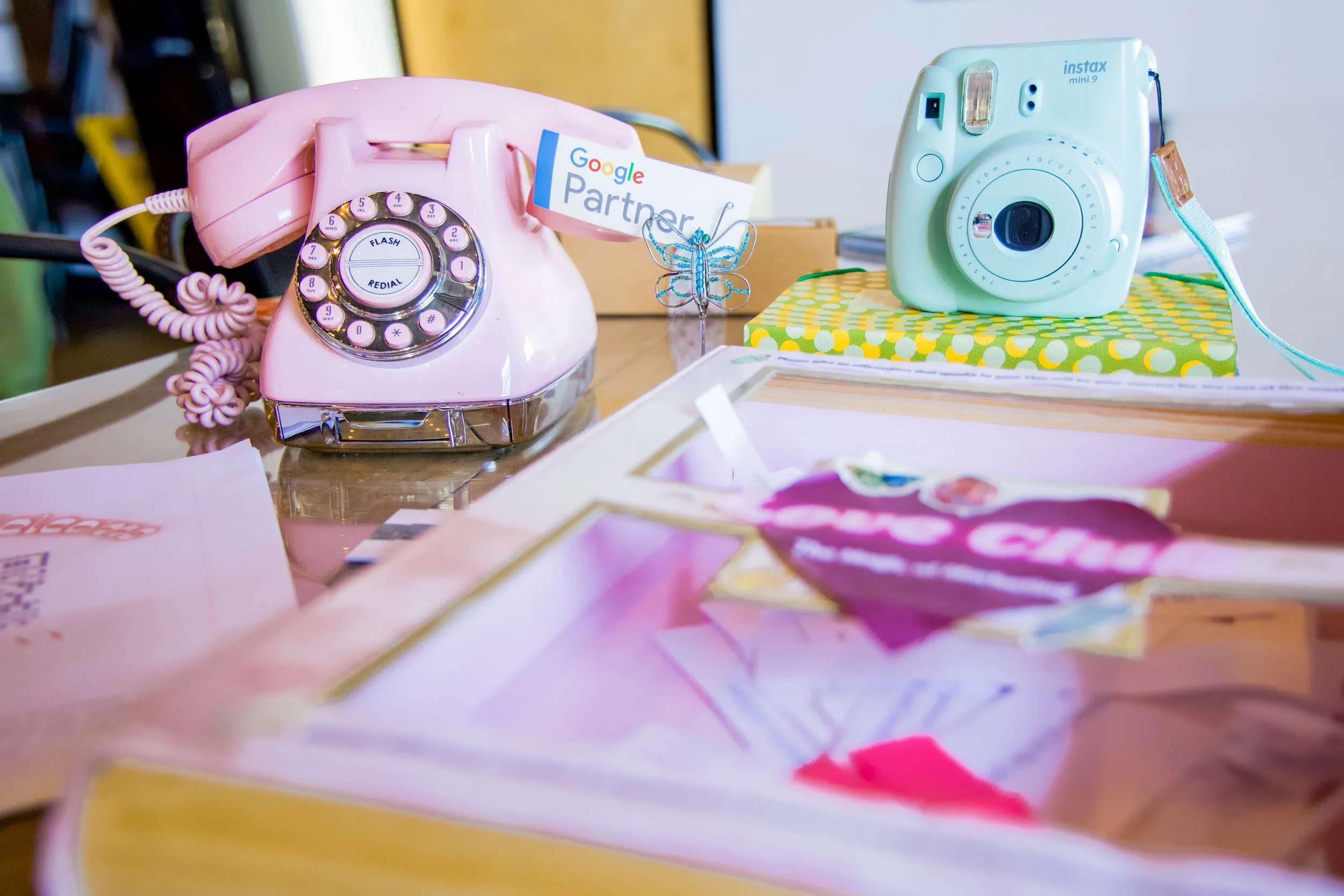 A pink rotary telephone and a mint green instant camera placed on a desk with various papers and objects.