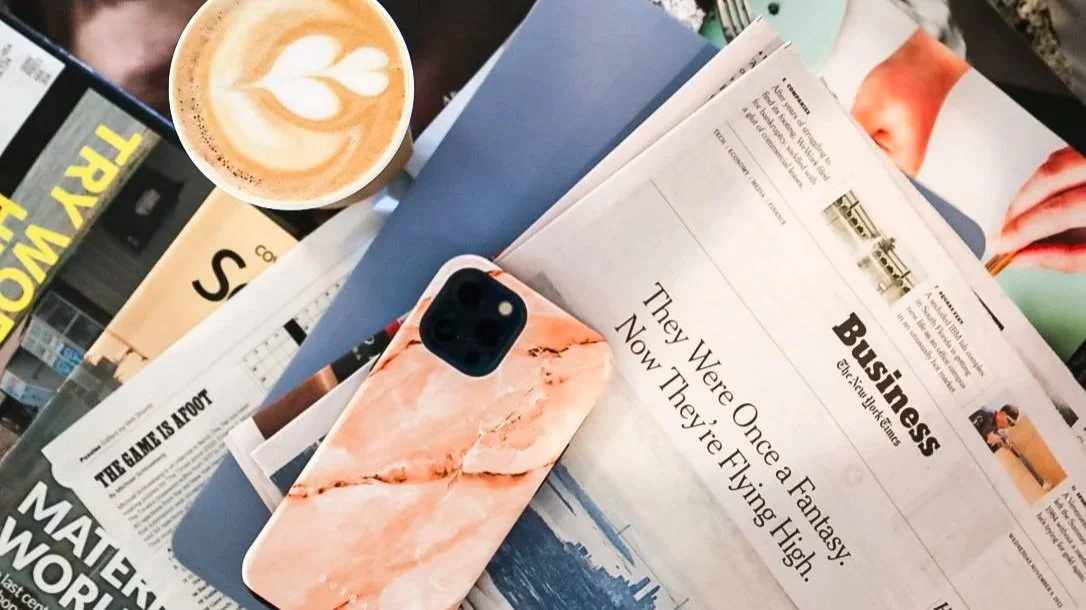 A top-down view of a workspace with a cup of coffee with foam art, a smartphone with a pink marble case, several open newspapers, and magazines on a wooden surface.