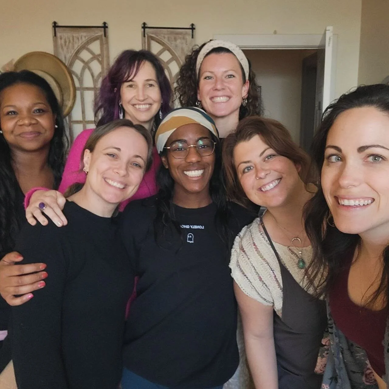 Group of eight women smiling and posing together indoors, some wearing jewelry and headbands, with a decorative wooden wall hanging in the background.