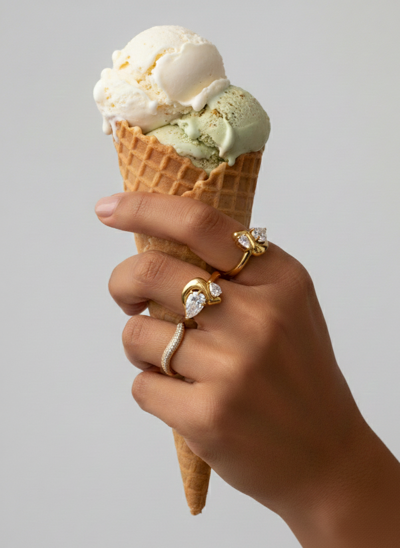 A person's hand holding a waffle cone with two scoops of ice cream, one vanilla and one pistachio, against a plain background. The person is wearing multiple gold rings with diamond accents.