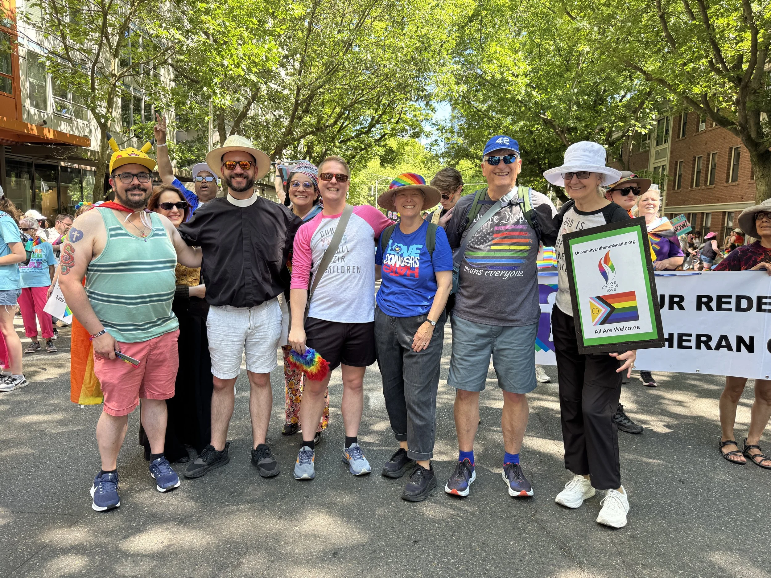 Group of people at a pride parade, smiling and posing for a photo, some wearing rainbow accessories, standing on a city street lined with trees and buildings, holding a rainbow flag and a sign.