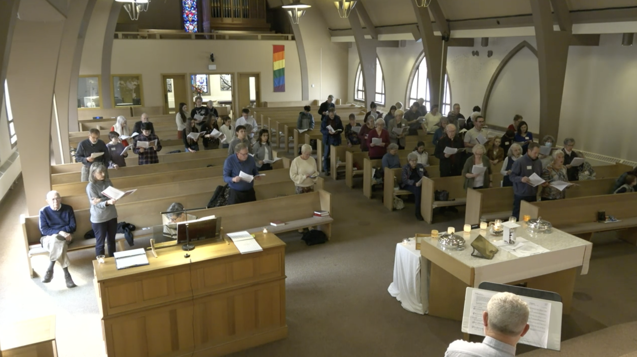 People gathered inside a church sitting in wooden pews, participating in what appears to be a religious service or prayer session. Some are holding hymn books or papers while standing or sitting.