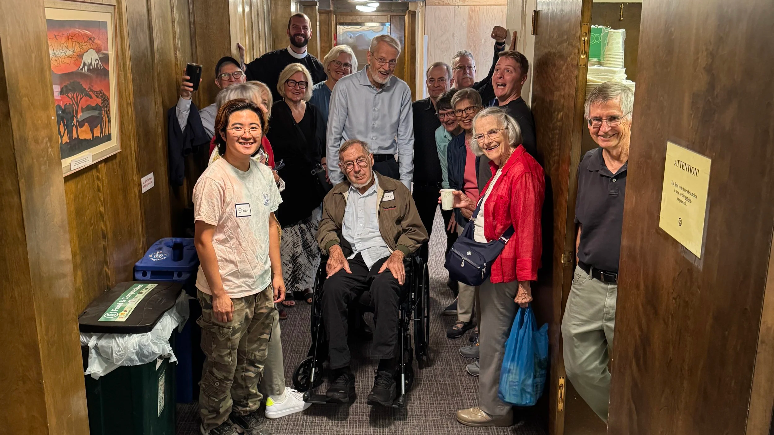 Group of smiling people, including an elderly man in a wheelchair, posing in a hallway with wooden walls decorated with artwork, celebrating at a social gathering.