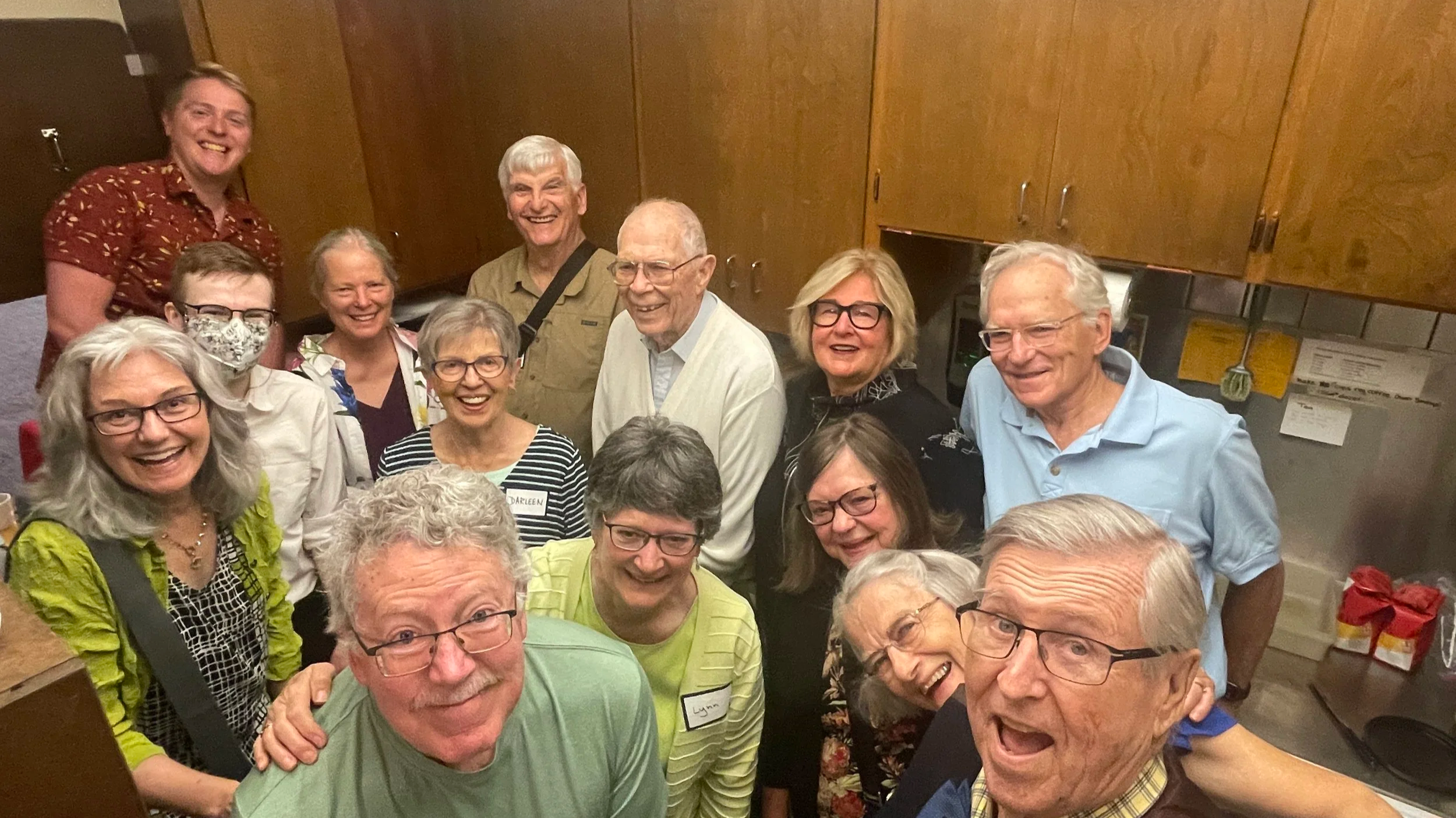 A group of fifteen smiling older adults gathered in a kitchen, some wearing glasses and name tags, celebrating together.