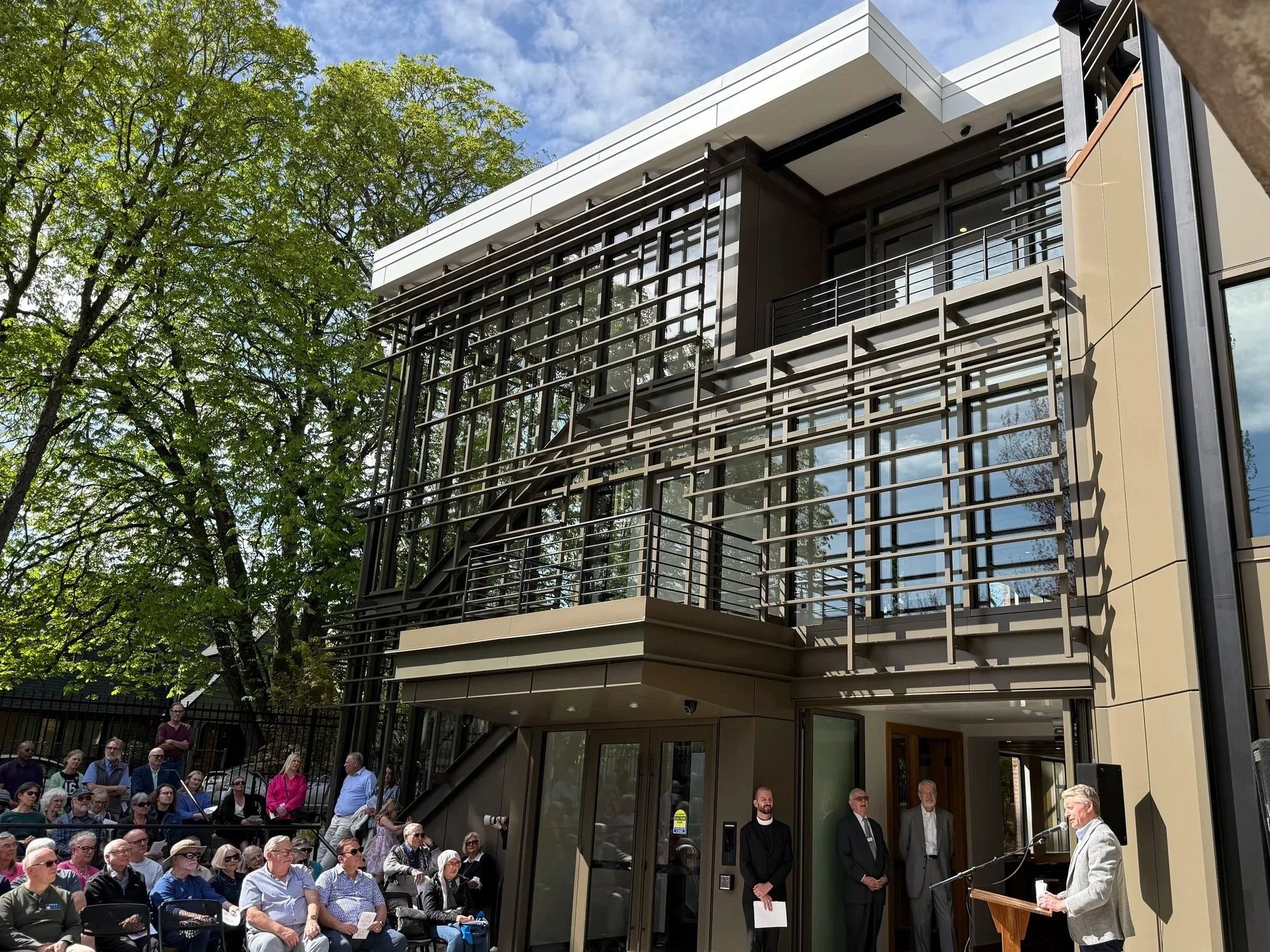A group of people seated outdoors in front of a modern building with glass windows and tree in the background, attending an event with a speaker at a podium.