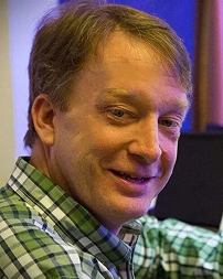 A man with light brown hair and a plaid shirt smiling at the camera in an indoor setting.
