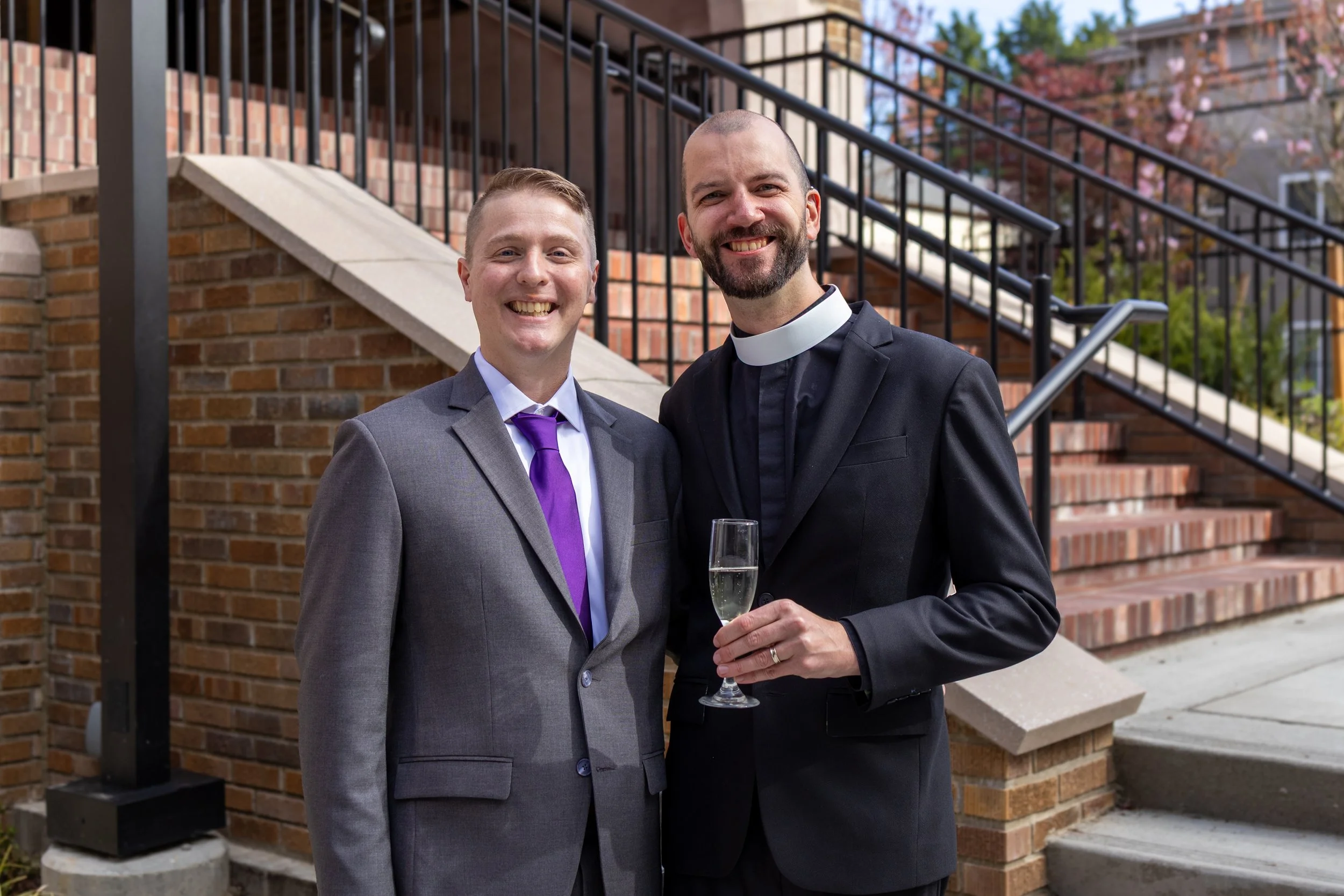 Two men in suits, one with a clerical collar, smiling and holding a glass of champagne outdoors in front of a brick building with stairs and a black iron railing, trees with pink blossoms in the background.