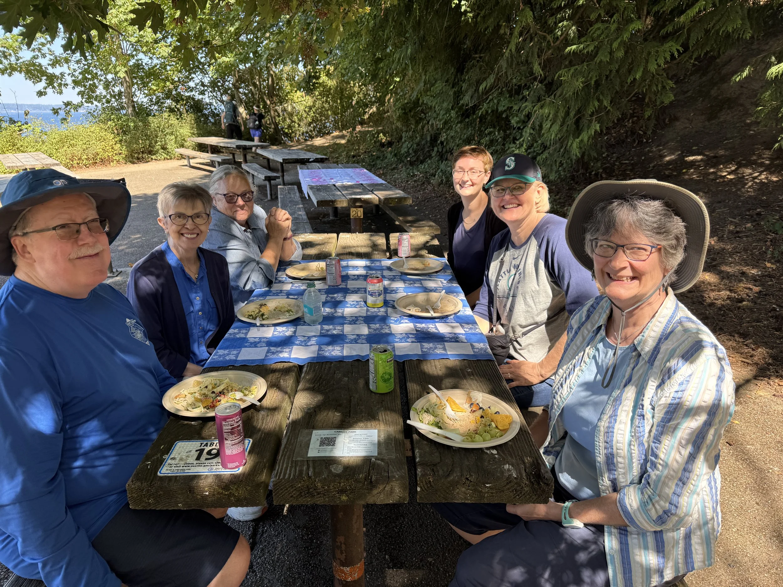 Group of seven people sitting at a picnic table outdoors, enjoying a meal on a sunny day surrounded by trees.