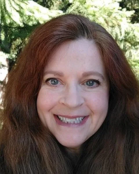 A woman with long red hair smiling outdoors with green foliage in the background.