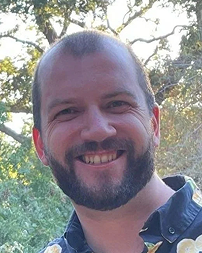Close-up of a smiling man with short hair and a beard outdoors, trees in the background.