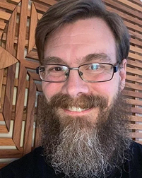 Close-up of a smiling man with glasses and a beard, with a wooden background.