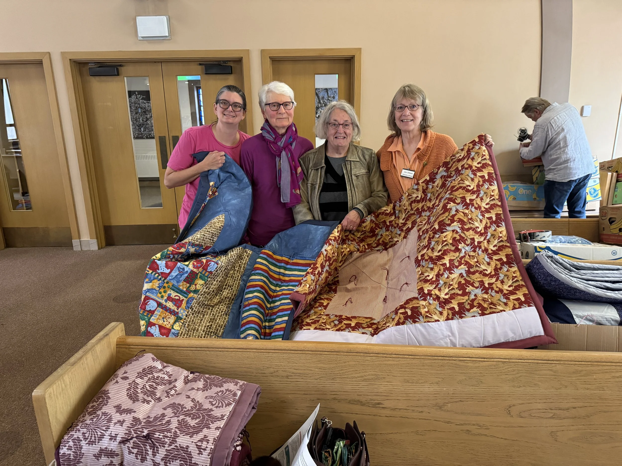 Four women standing together indoors in front of a wooden door, holding a pile of colorful folded blankets or quilts, with a man in the background looking into a box.