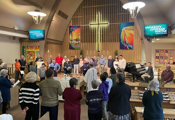 A group of people gathered in a church or community hall for a choir or musical performance. The setting features a cross, colorful banners, a piano, and two screens displaying the text 'University Lutheran Church of Seattle.'