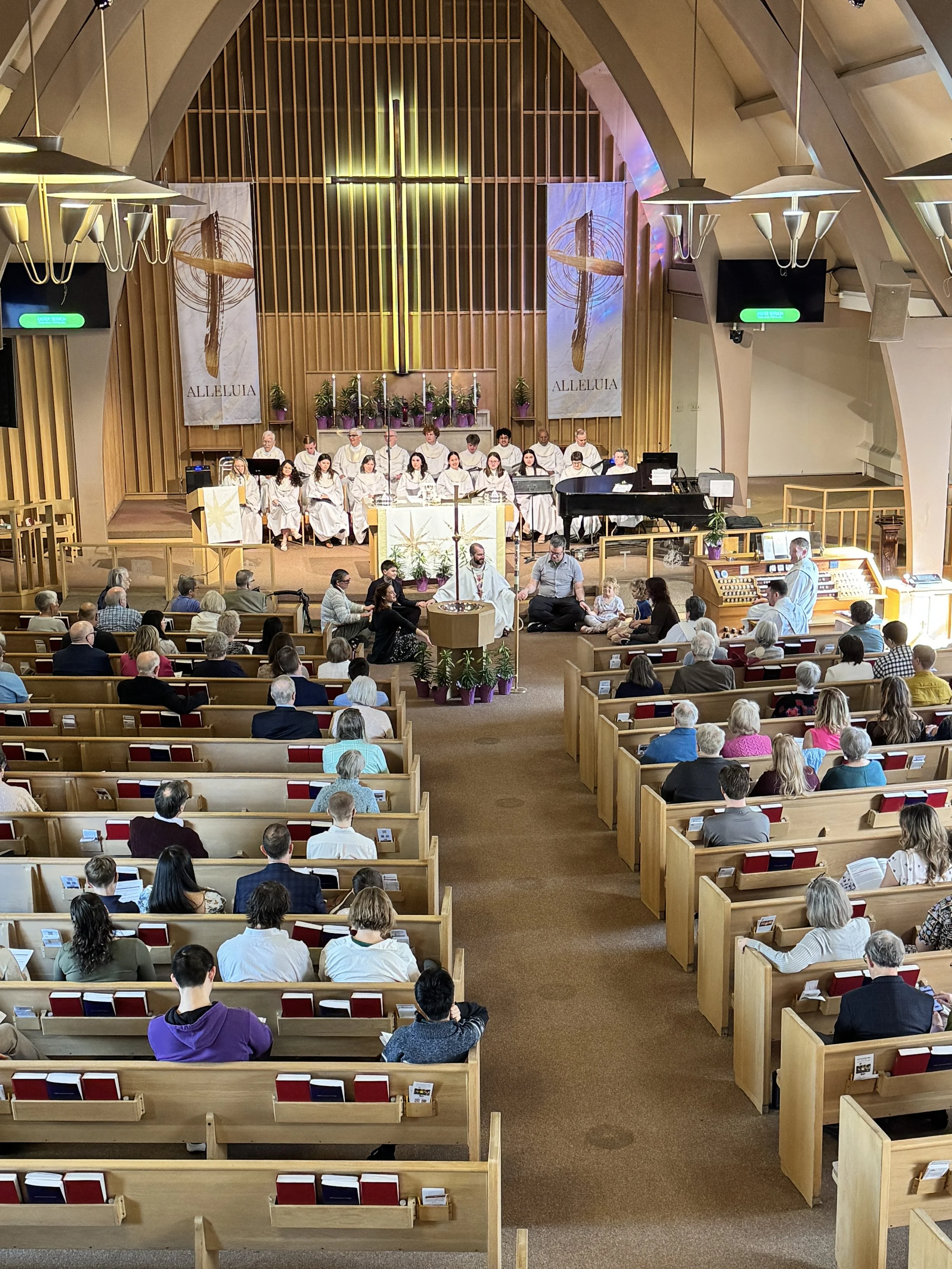 Inside a church with a congregation attending service, choir in white robes at the front, altar with candles and plants, wooden pews filled with people, wooden architecture, large cross on the wall, banners with religious imagery and the text 'ALLELUIA', and a piano.