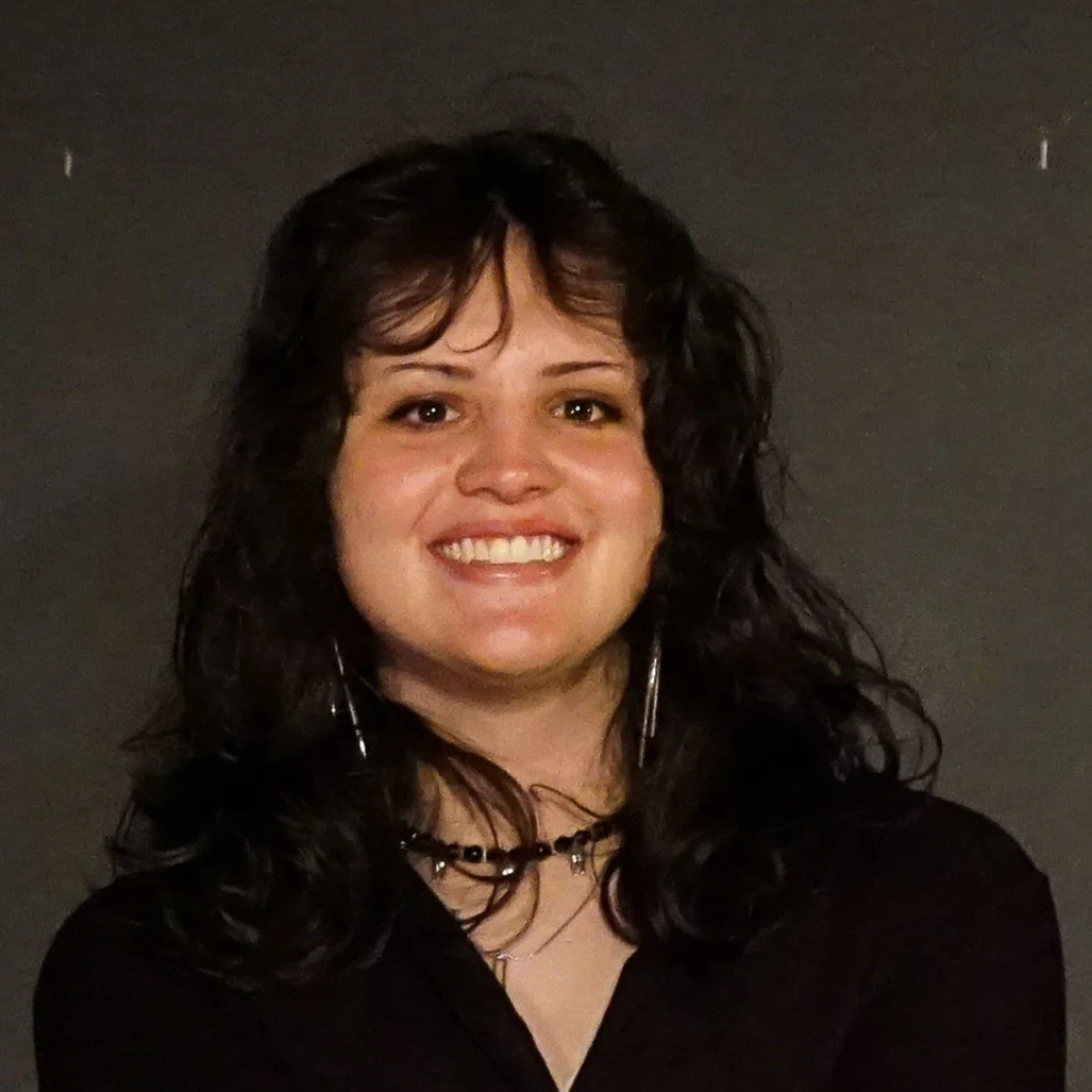 A woman with dark curly hair smiling in front of a plain dark background.