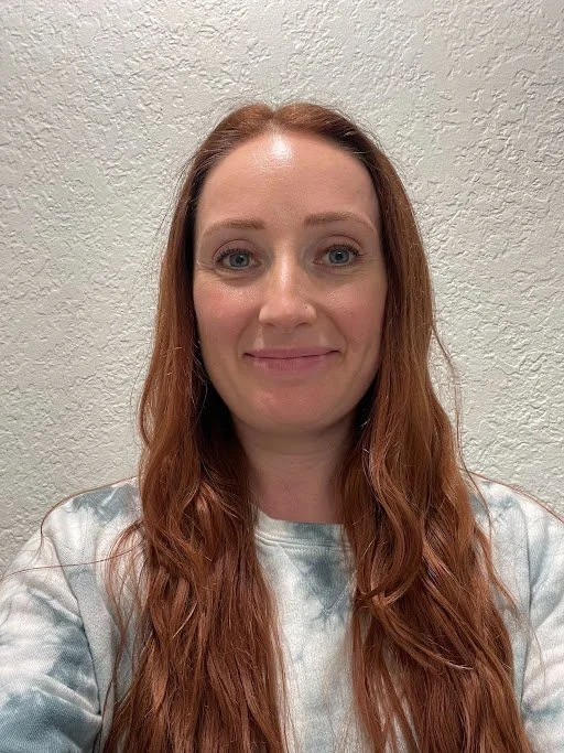 A woman with long red hair and blue eyes smiling at the camera in front of a textured white wall.