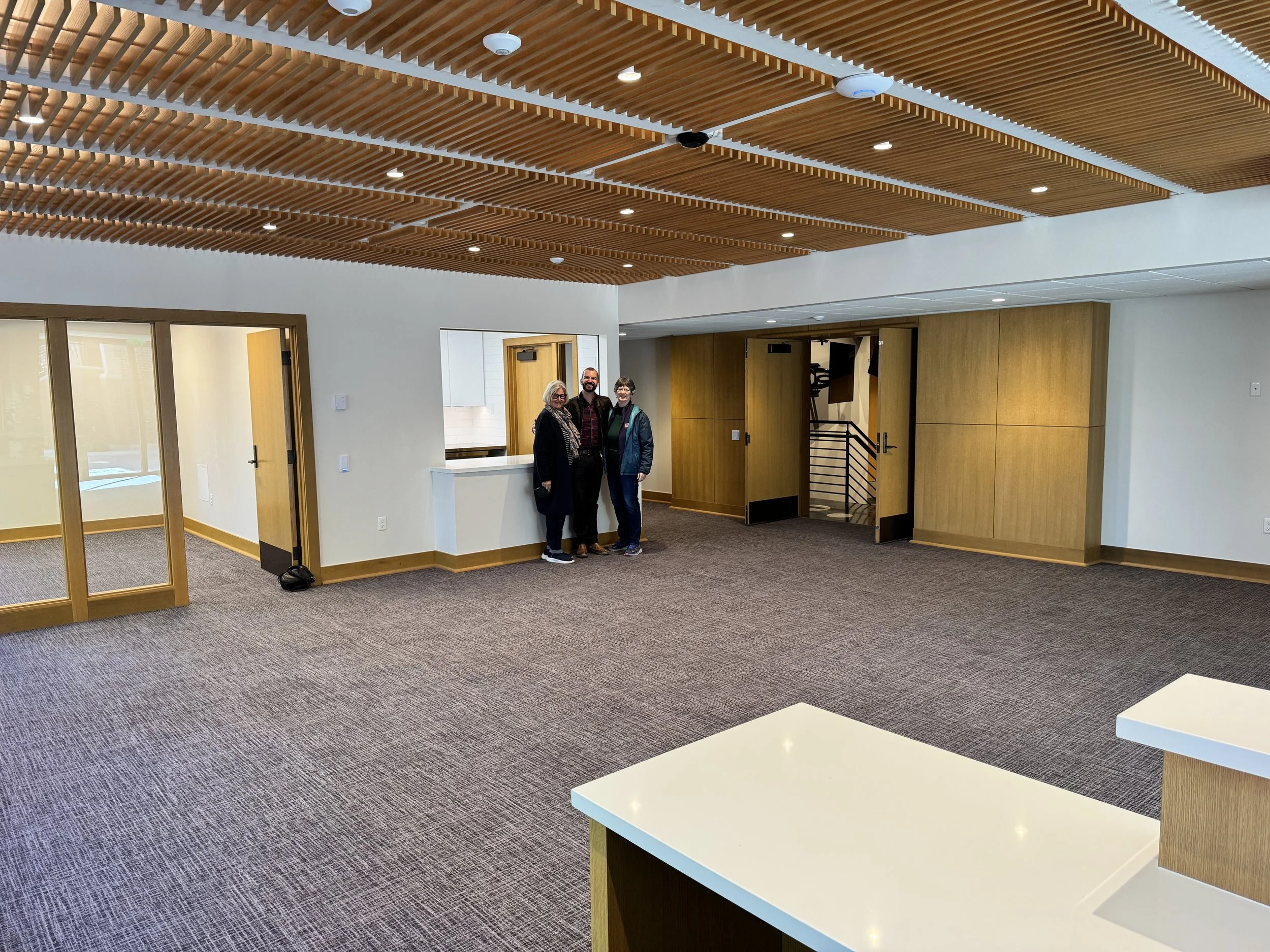 Interior of a modern office or conference room with wooden slatted ceiling, beige walls, and brown carpeting. Three people are standing near a white reception desk, smiling at the camera. There are glass doors and wooden partition walls in the room.