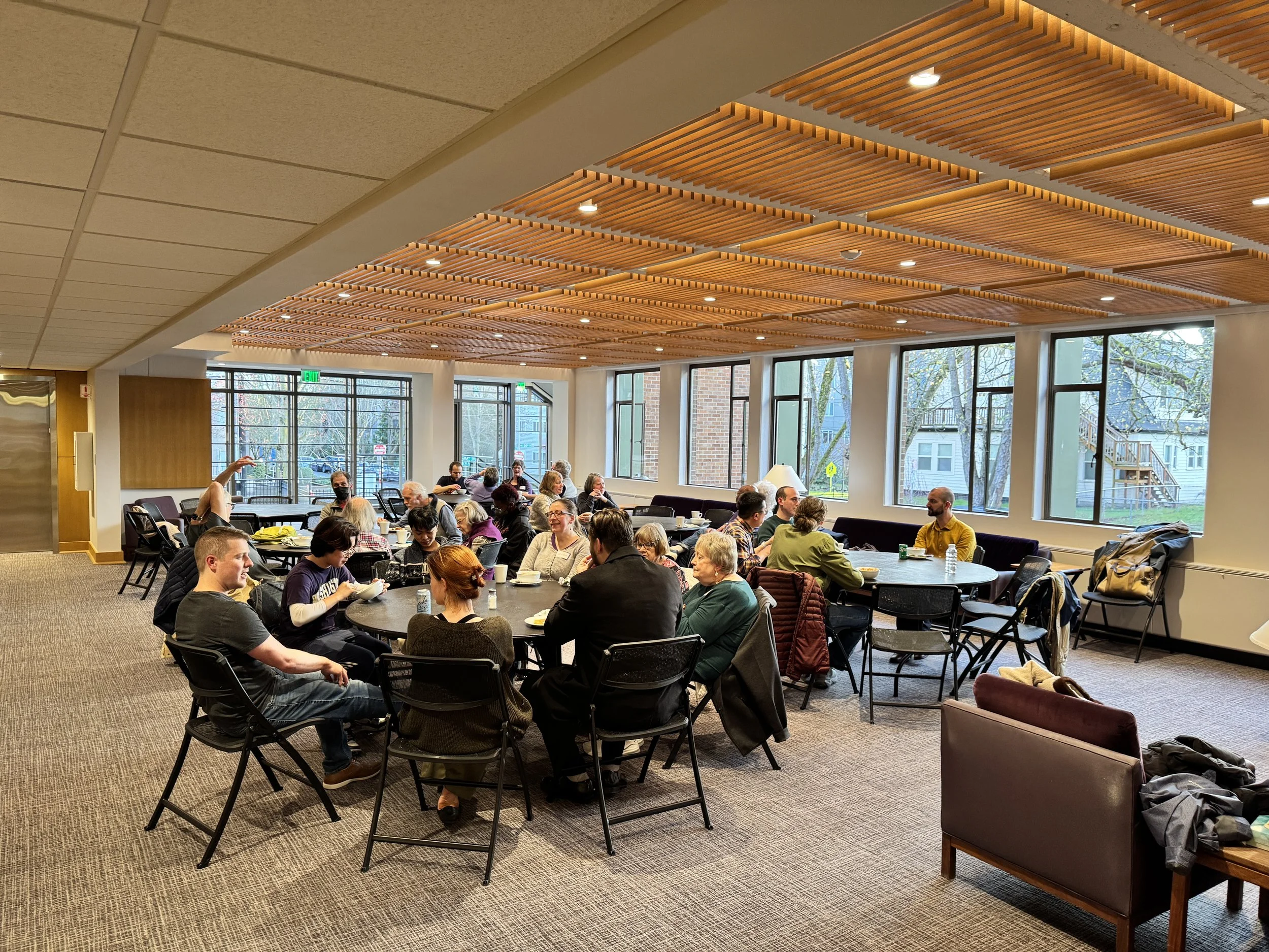 People sitting at tables in a bright room with large windows, wooden ceiling decor, and trees visible outside.