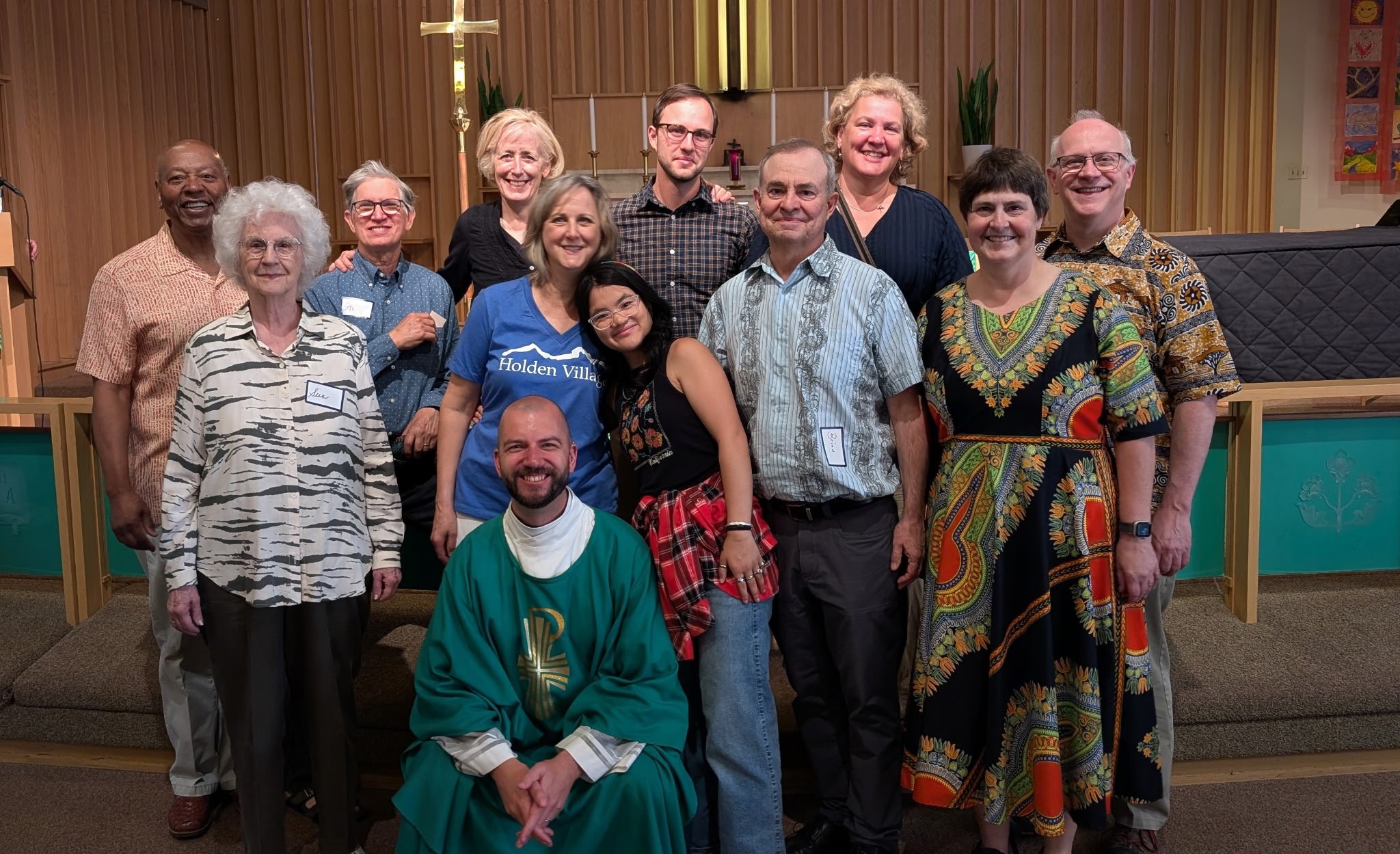 A group of fifteen diverse people gathered inside a church or community center, posing for a photo in front of a wooden altar with green plants and candles, smiling and dressed in casual and semi-formal attire.