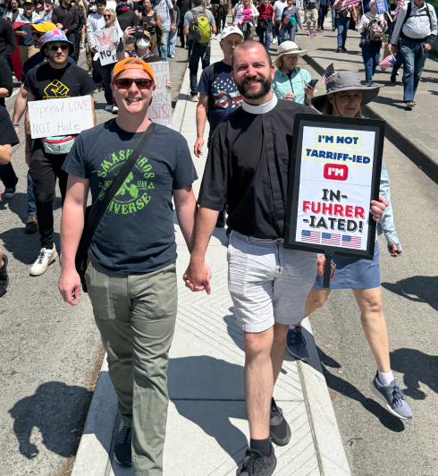 Two men holding hands and smiling at a public protest or march. One man is dressed as a clergy member or pastor and holding a sign that reads 'I'm not tariff-ified, I'm in-fuh-hrer-tated!' The other man is wearing sunglasses and a T-shirt.