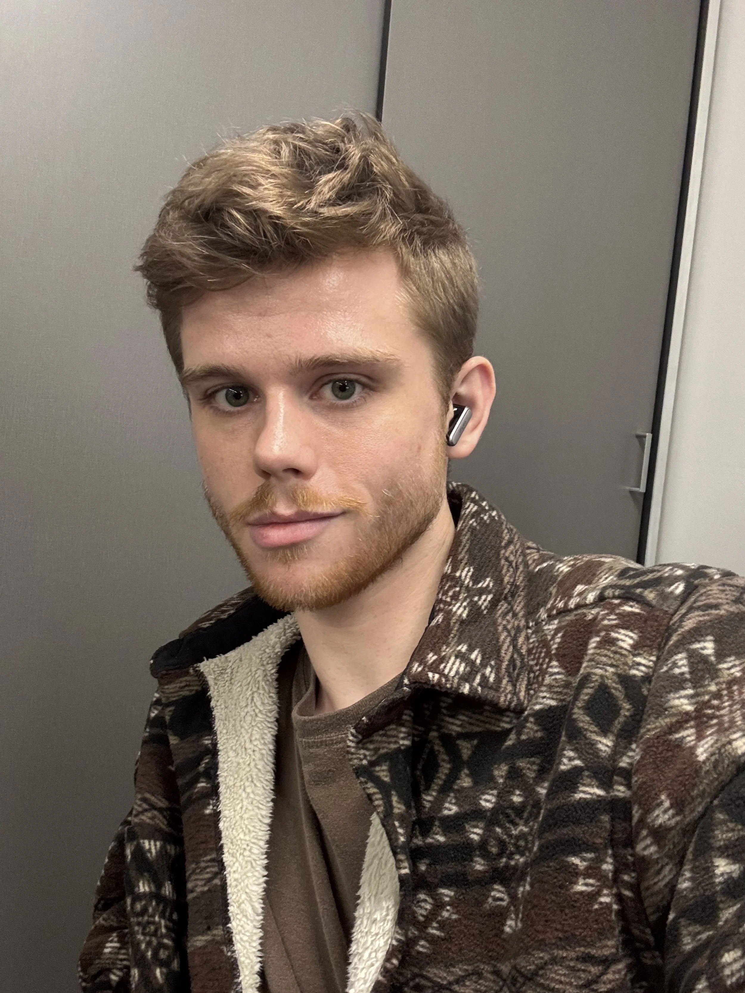 A young man with short, wavy, light brown hair and blue eyes taking a selfie. He's wearing a brown patterned jacket and a brown shirt underneath. He's also wearing a wireless earbud in his right ear. The background is a plain, gray wall.