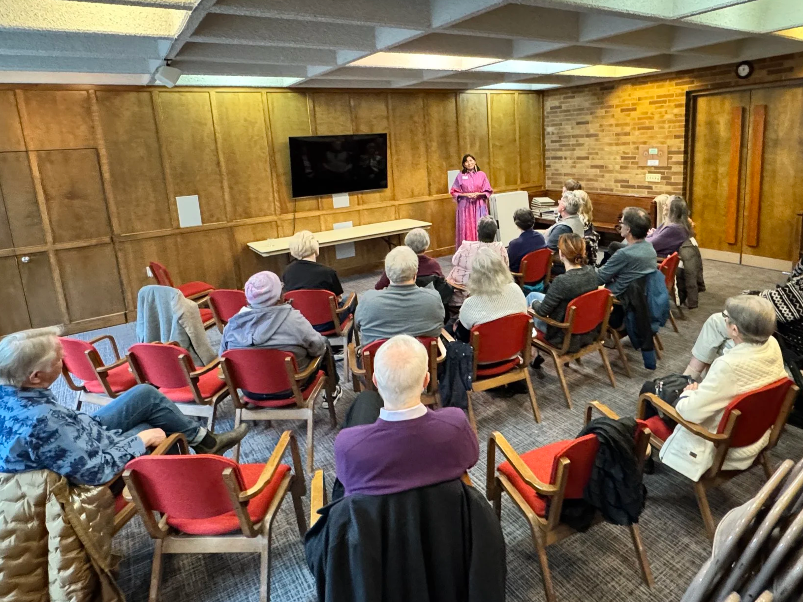 A woman in a pink dress is speaking to an audience seated in red chairs in a room with wood-paneled walls and a brick wall. There is a large TV screen on the wall behind her.