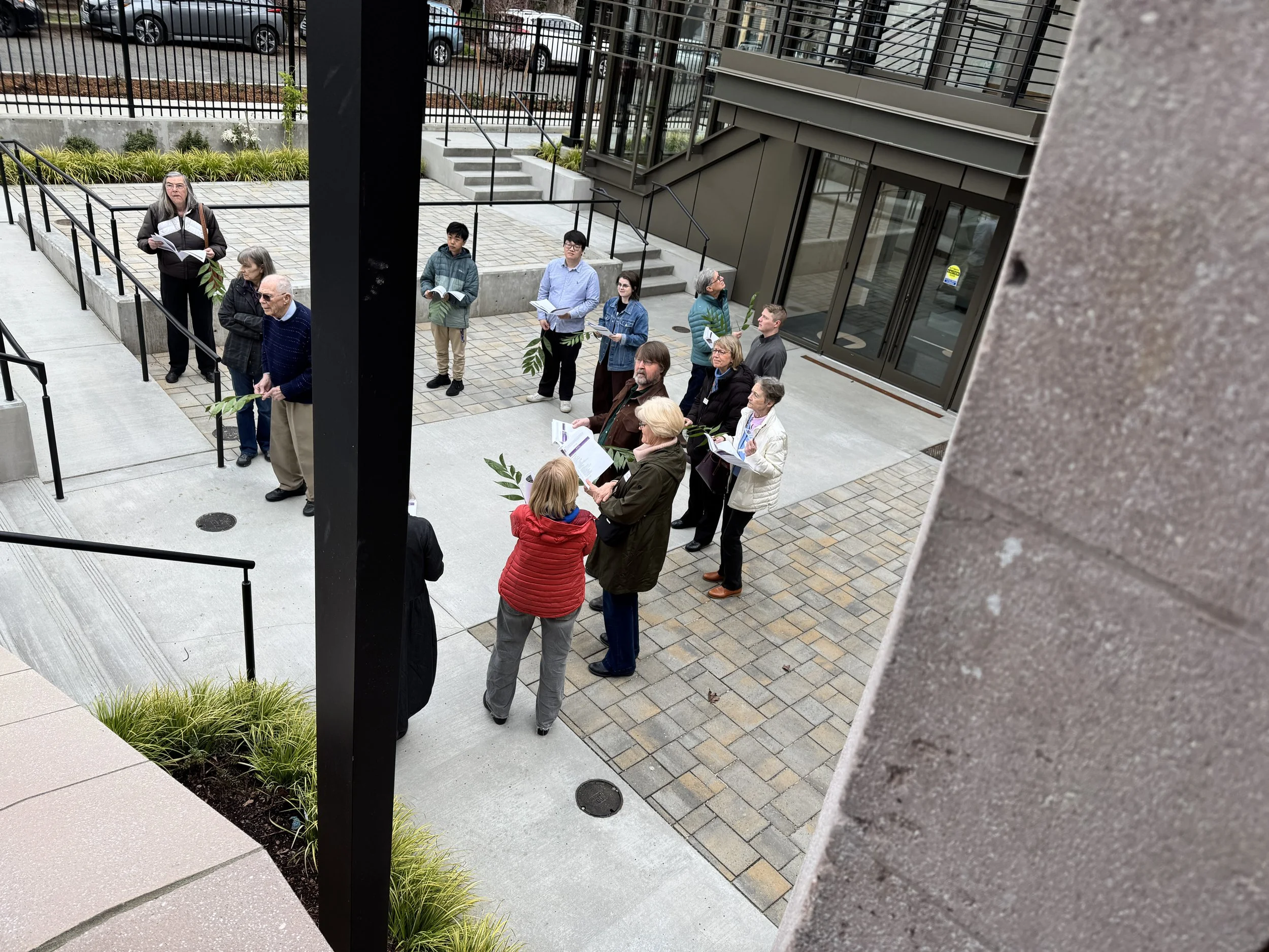 A group of diverse people standing in line outside a modern building, holding papers or booklets, possibly waiting for an event or tour, with stairs, glass doors, and a landscaped area with plants nearby.