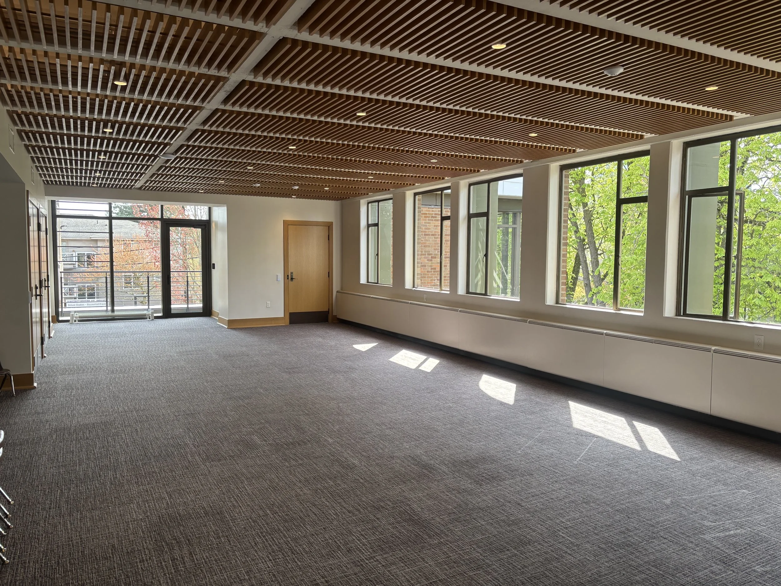 Empty indoor room with large windows, wooden ceiling panels, and a grey carpeted floor, sunlight streaming in from outside.