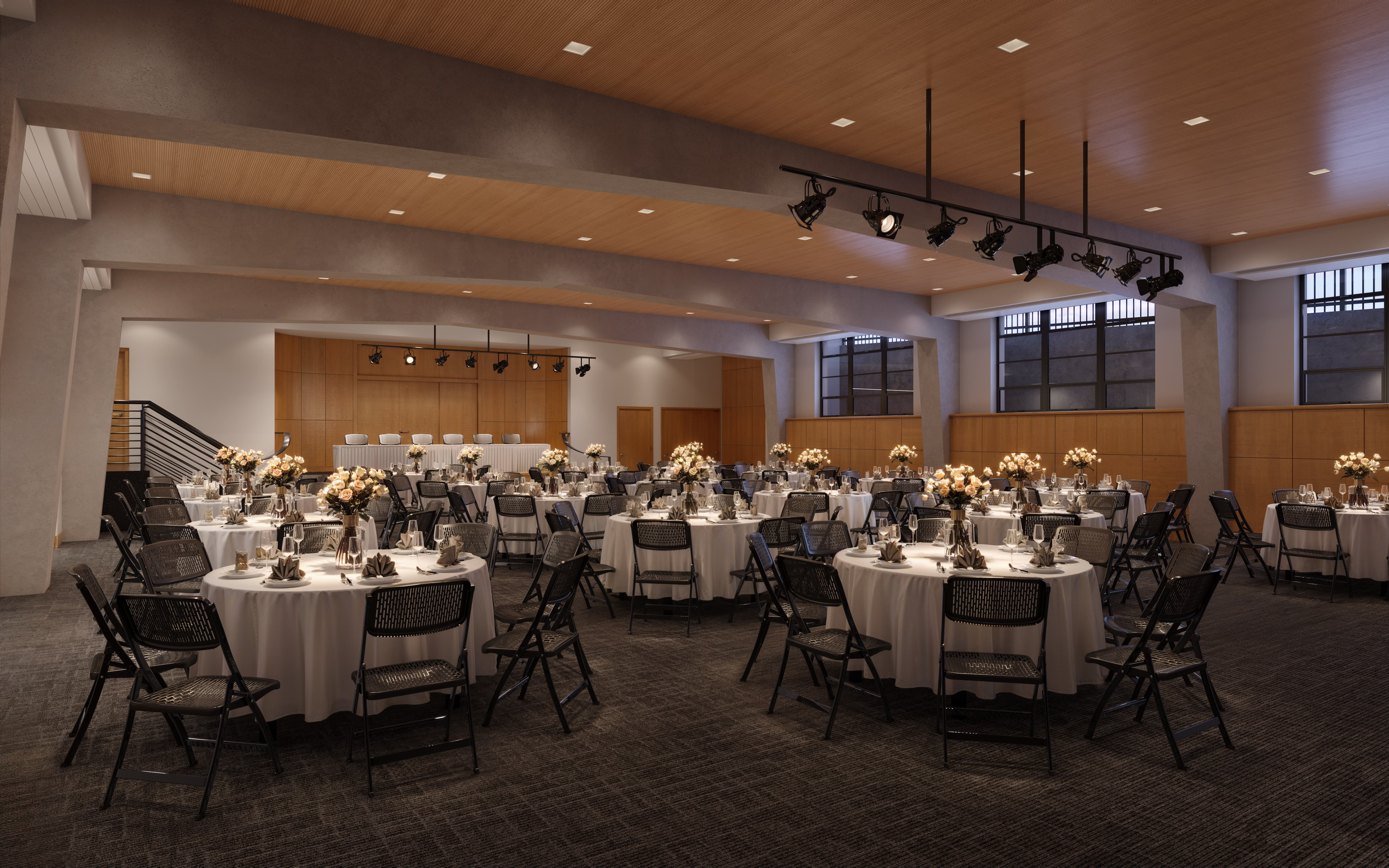 Banquet hall decorated for an event with round tables covered with white tablecloths, floral centerpieces, and place settings. There are large windows, a stage area in the distance, and professional lighting equipment hanging from the ceiling.