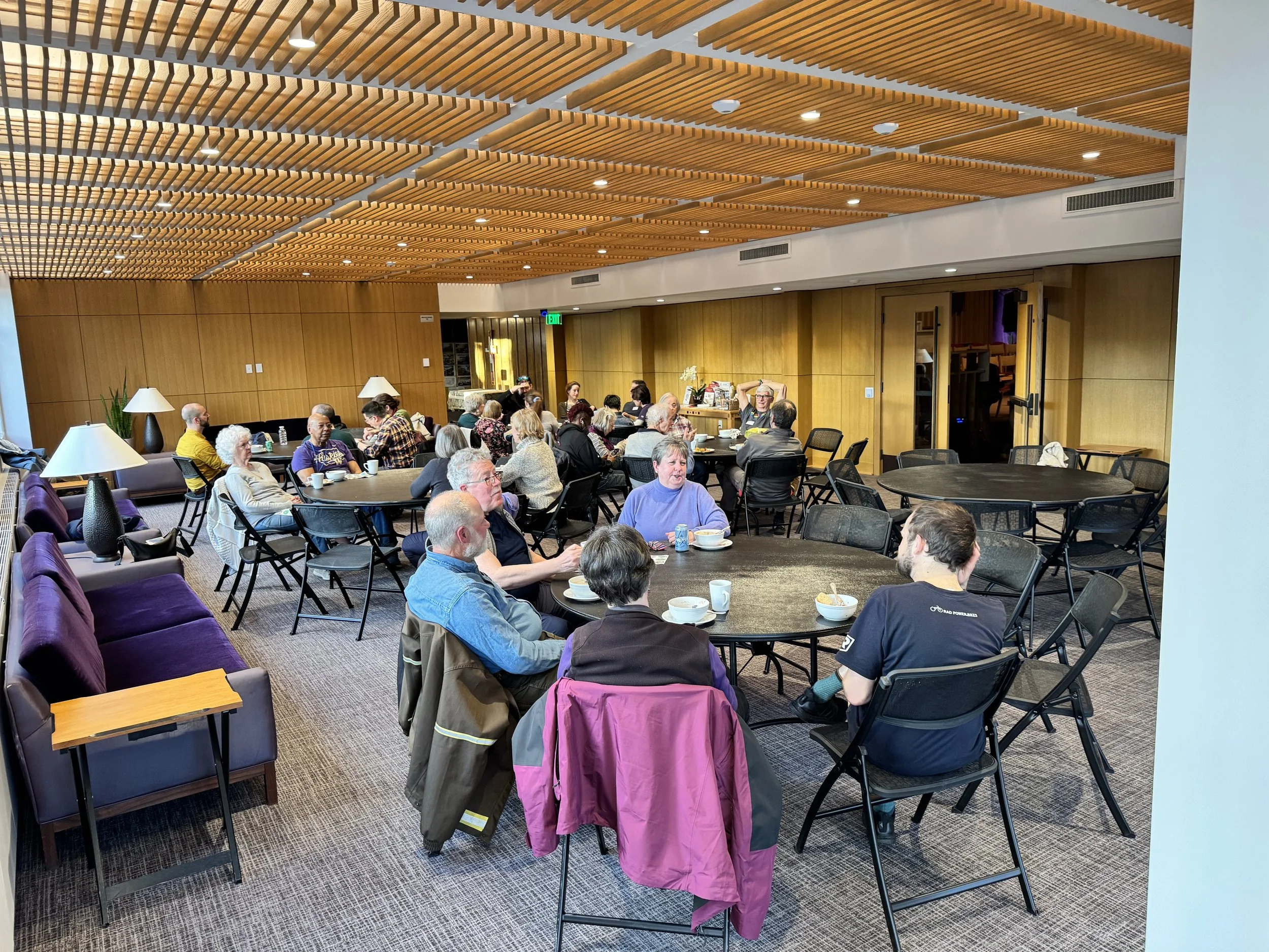 A group of people gathered in a modern indoor lounge area, sitting at round tables, some eating or drinking, with warm wooden wall and ceiling decor.