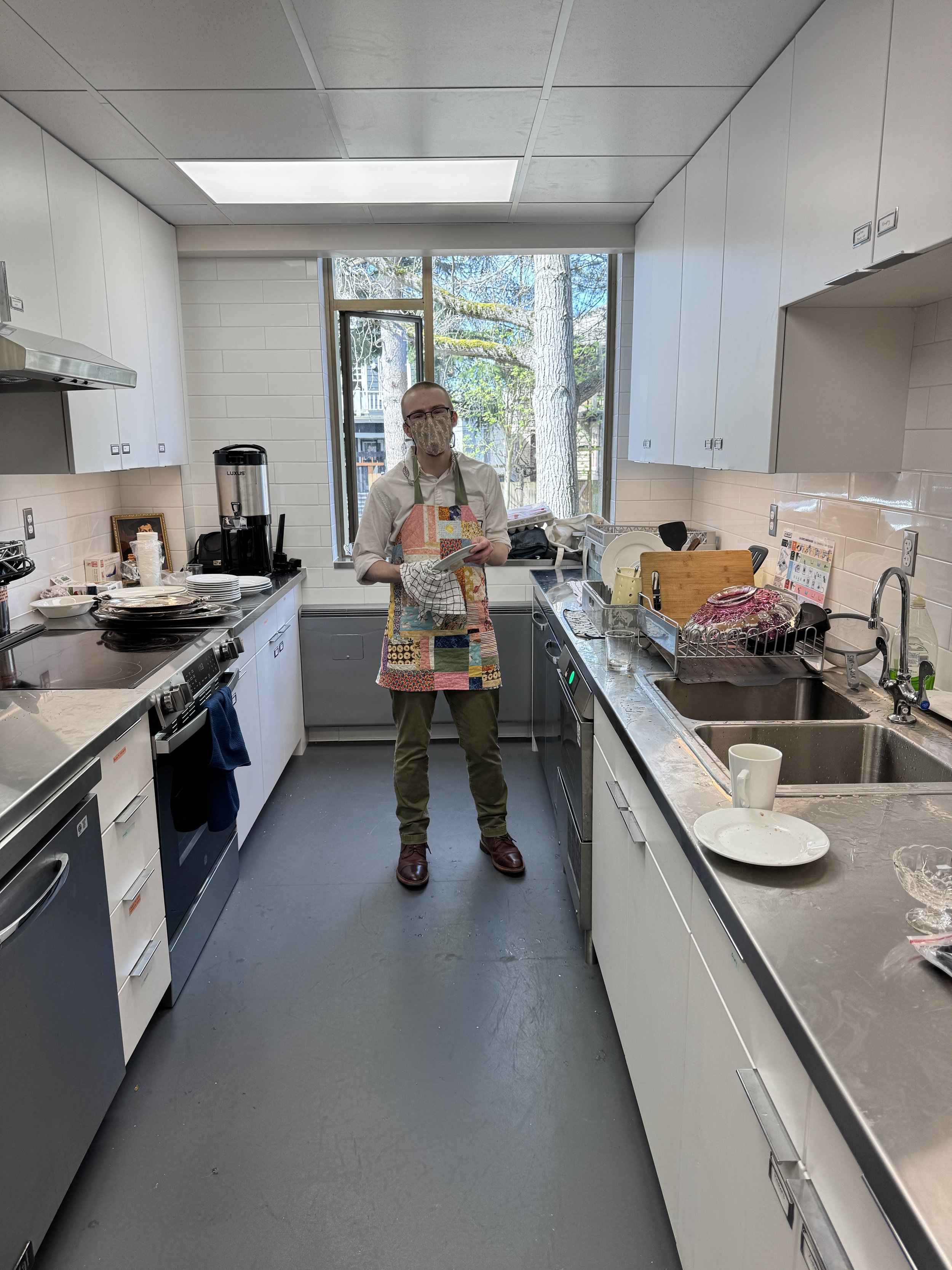 A man wearing glasses, a cloth face mask, a white shirt, and a colorful apron stands in a modern kitchen, holding a cloth in one hand. The kitchen features white cabinets, a double sink, a coffee machine, and various dishes and utensils on the countertops. Large windows in the background reveal trees outside.