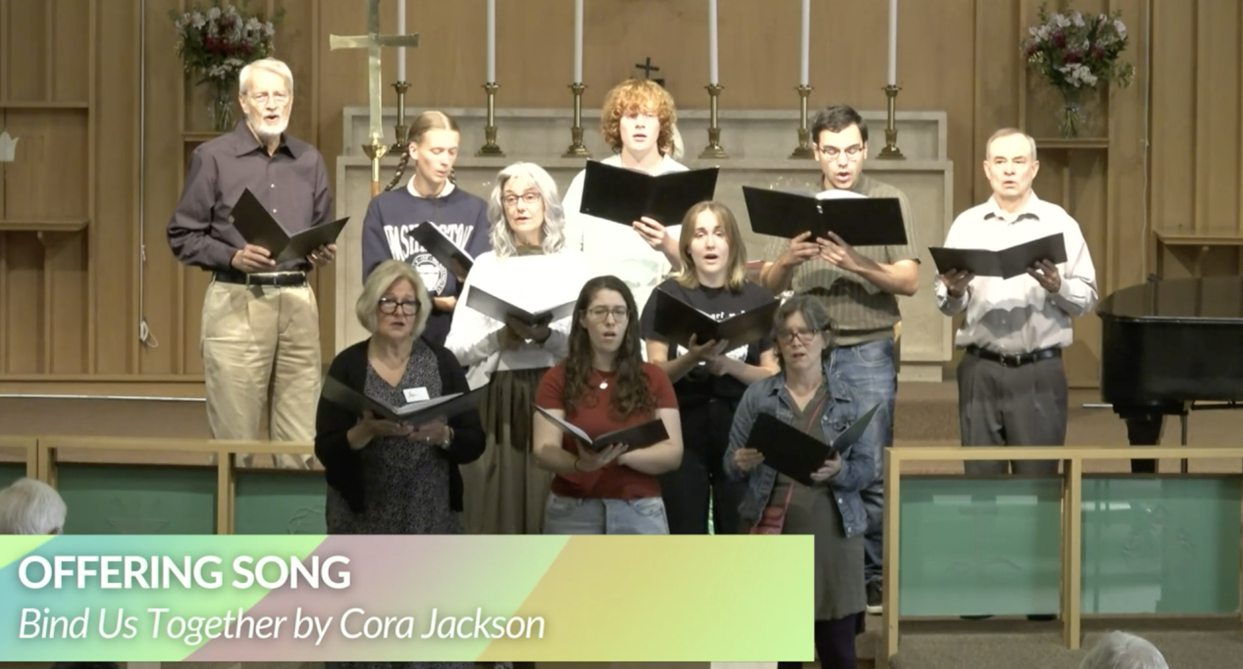 A group of people singing in a church choir with some holding black songbooks, set against a wooden background with candles and a cross. Text overlay reads 'OFFERING SONG' and 'Bind Us Together by Cora Jackson'.