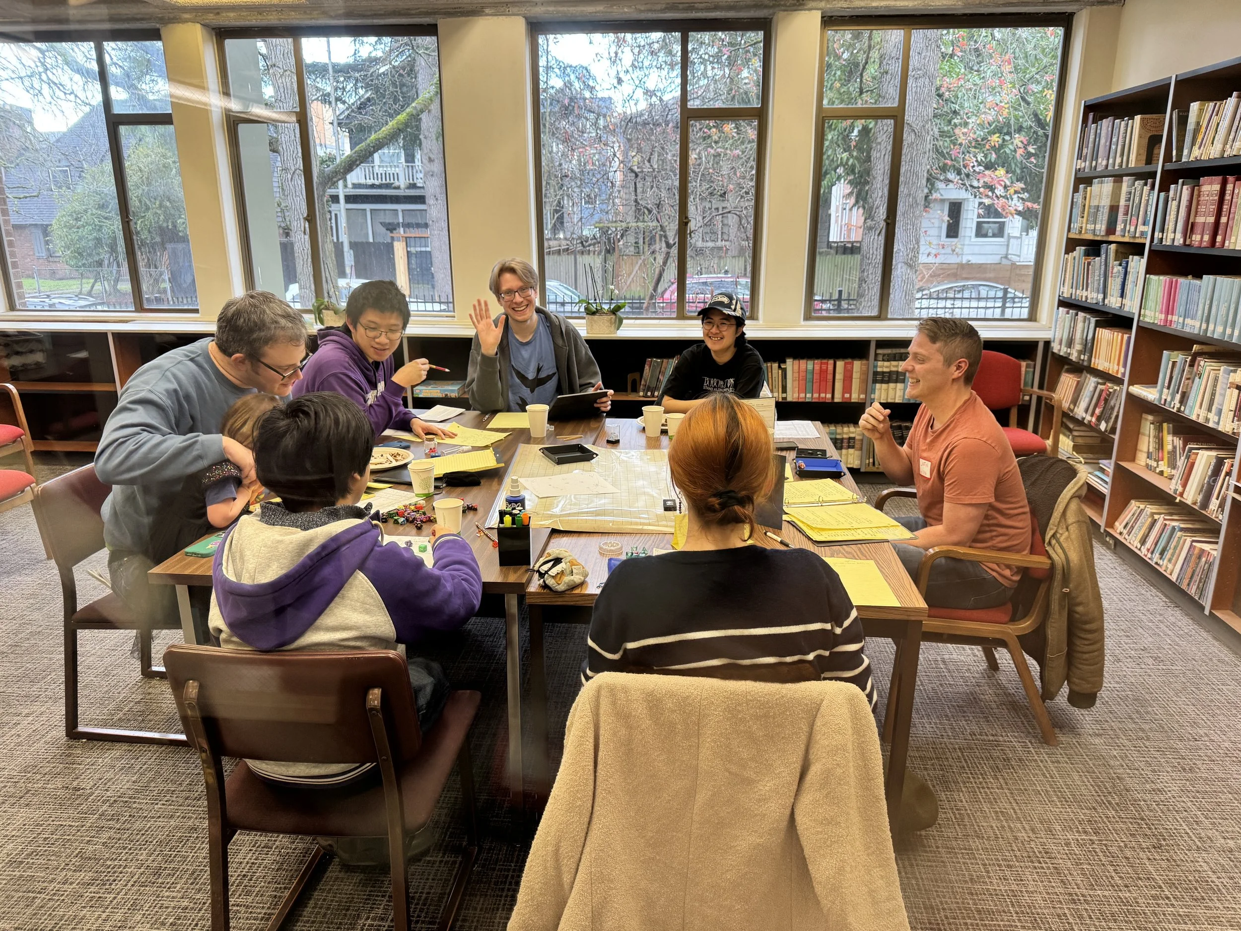 A diverse group of people gathered around a table in a library or community room, engaged in a discussion or game. The room has large windows showing trees and houses outside, and bookshelves filled with books are visible.