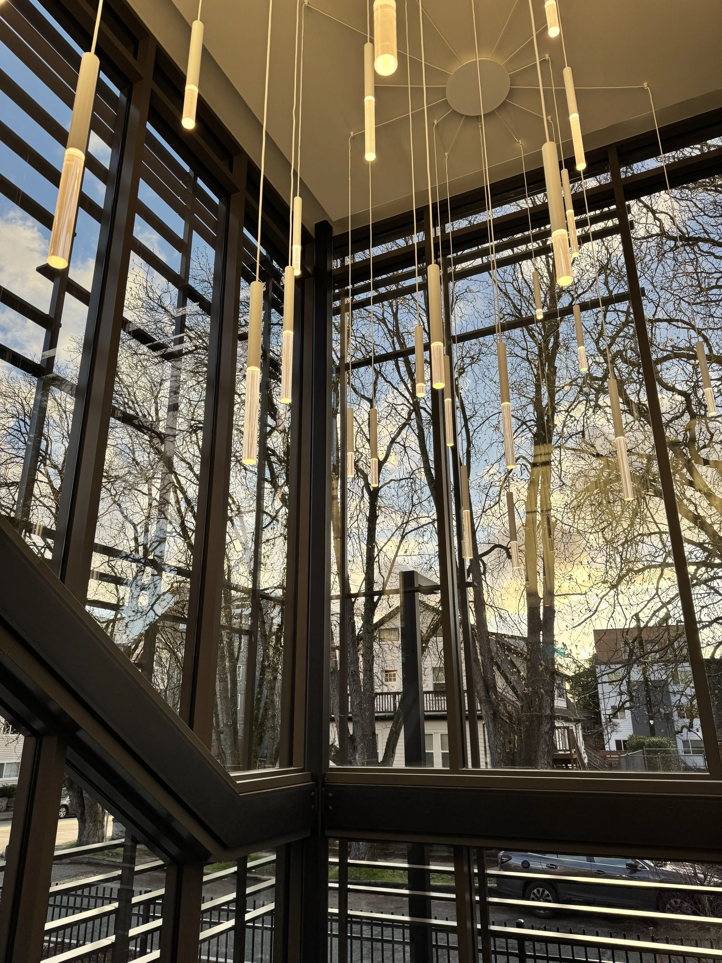 Interior view of a modern building with large glass windows showing trees and buildings outside. A contemporary chandelier with multiple hanging cylindrical lights is attached to the ceiling.
