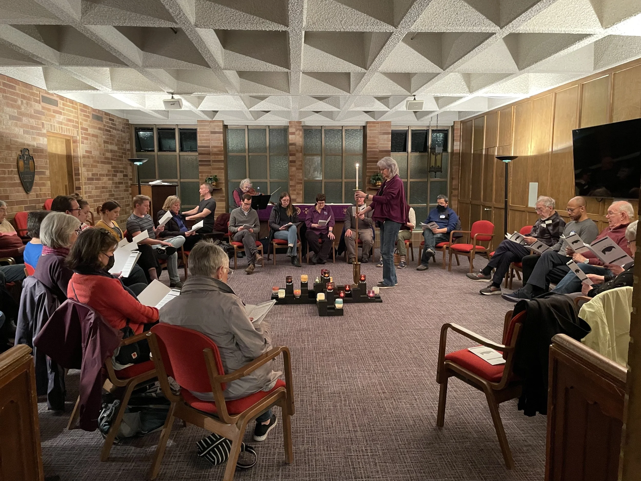 A group of people sitting in a circle in a room with brick and wood-paneled walls, participating in a spiritual or religious ceremony. One woman is standing and holding a lit candle, leading the session. Candles are arranged on the floor in front of the group, creating a peaceful atmosphere.