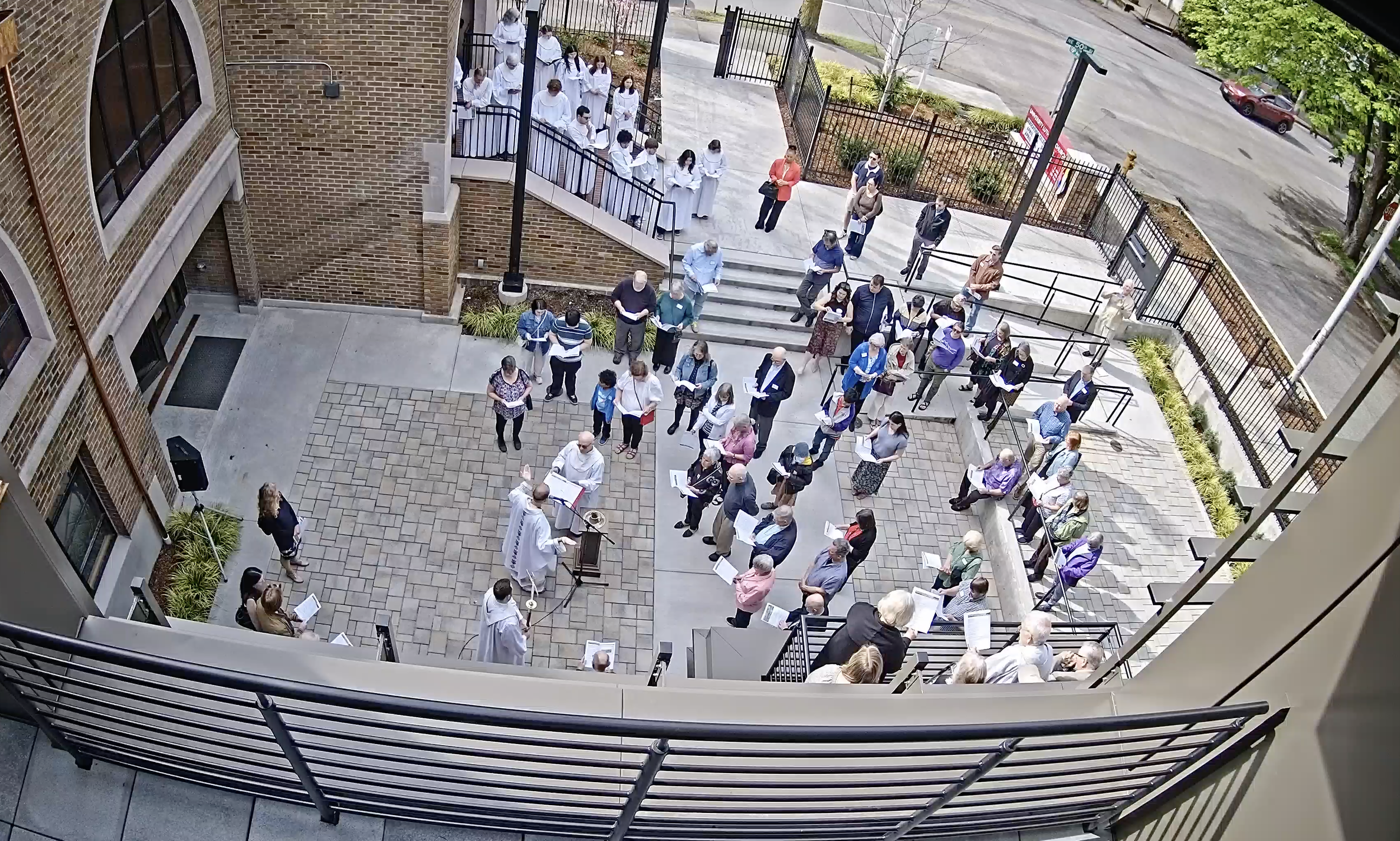 People gathered outside a brick building and sitting on the stairs, attending a religious service with a priest standing at a lectern.
