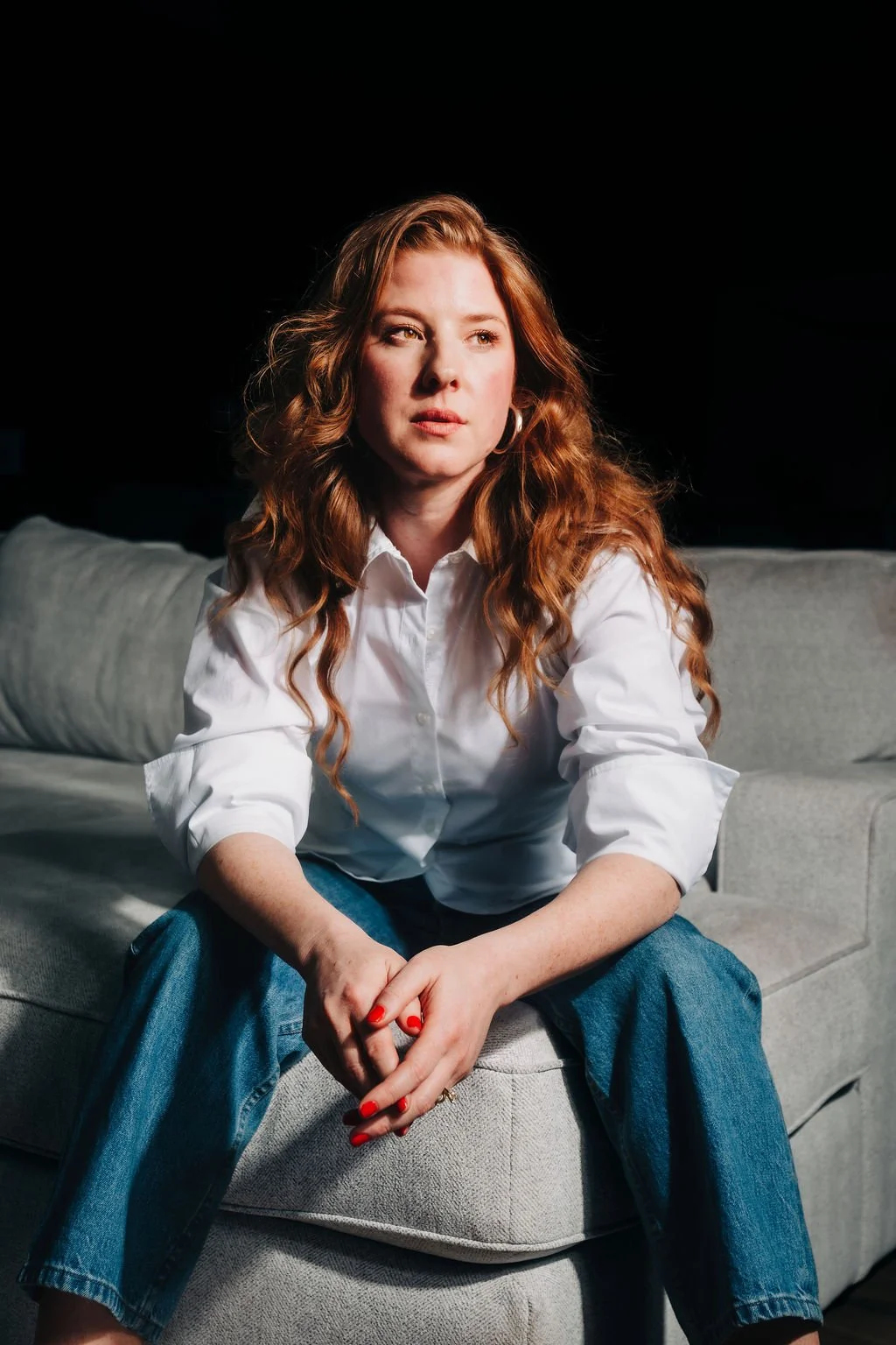 A woman trauma therapist with wavy red hair, wearing a white shirt and blue jeans, sitting on a light gray couch in a dark room, looking off to the side.