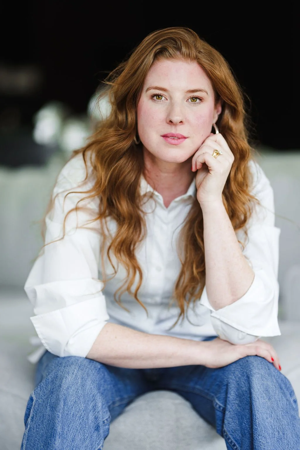 A woman trauma therapist with long red hair wearing a white shirt and blue jeans, sitting on a light-colored sofa with her hand resting on her face, looking directly at the camera.