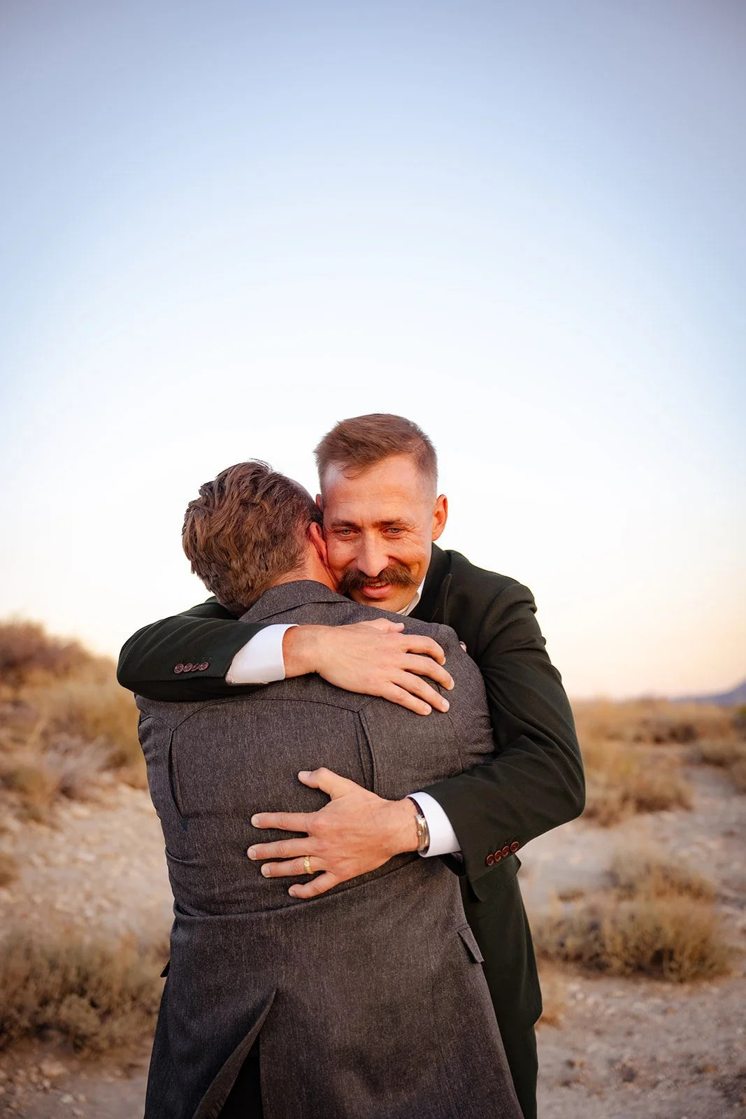 Two men in suits embrace outdoors during sunset, with a clear sky and desert landscape in the background.