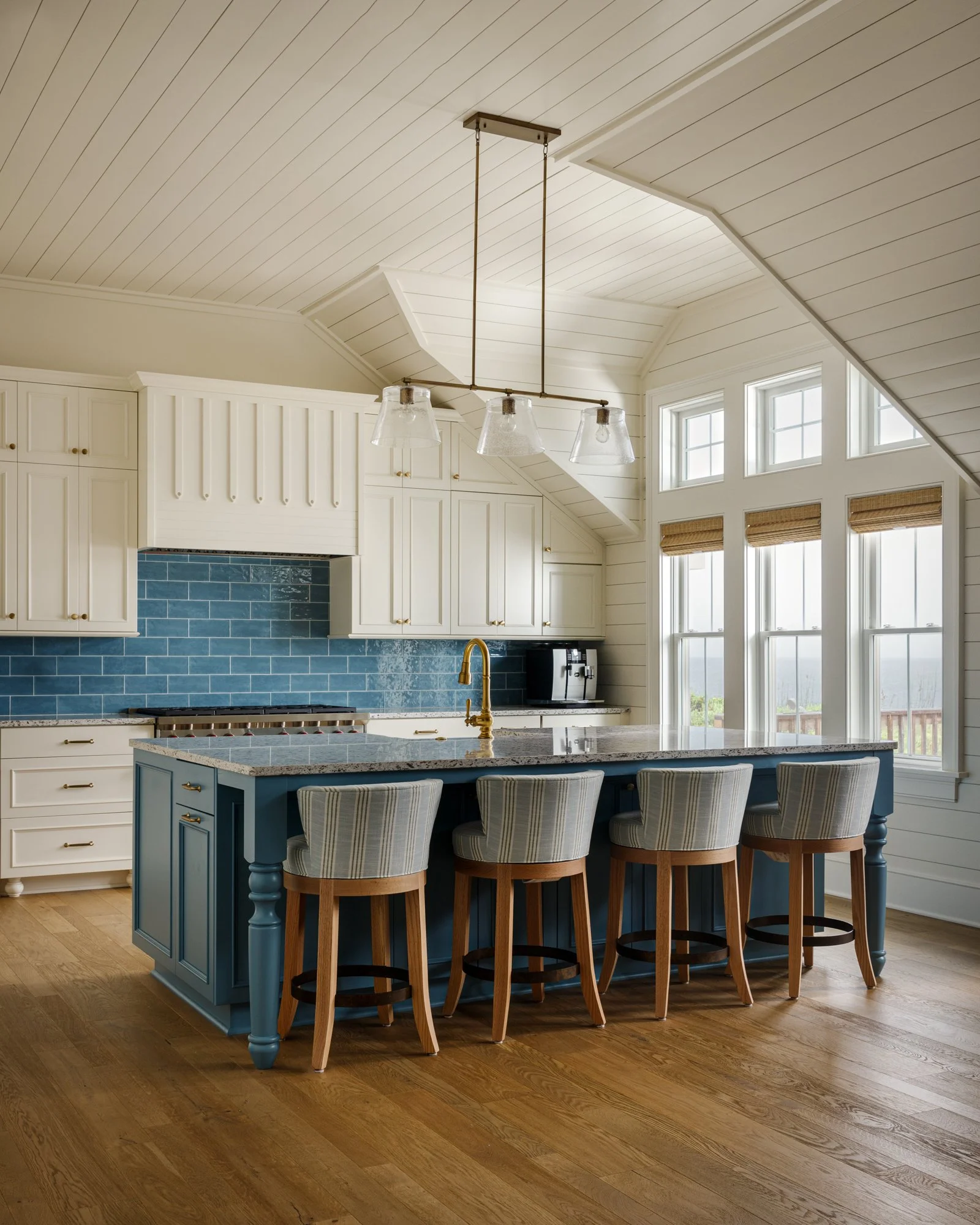 Modern kitchen with white cabinets, blue backsplash, island with blue base and granite countertop, four striped upholstered barstools, hardwood floor, and large windows with wooden blinds.