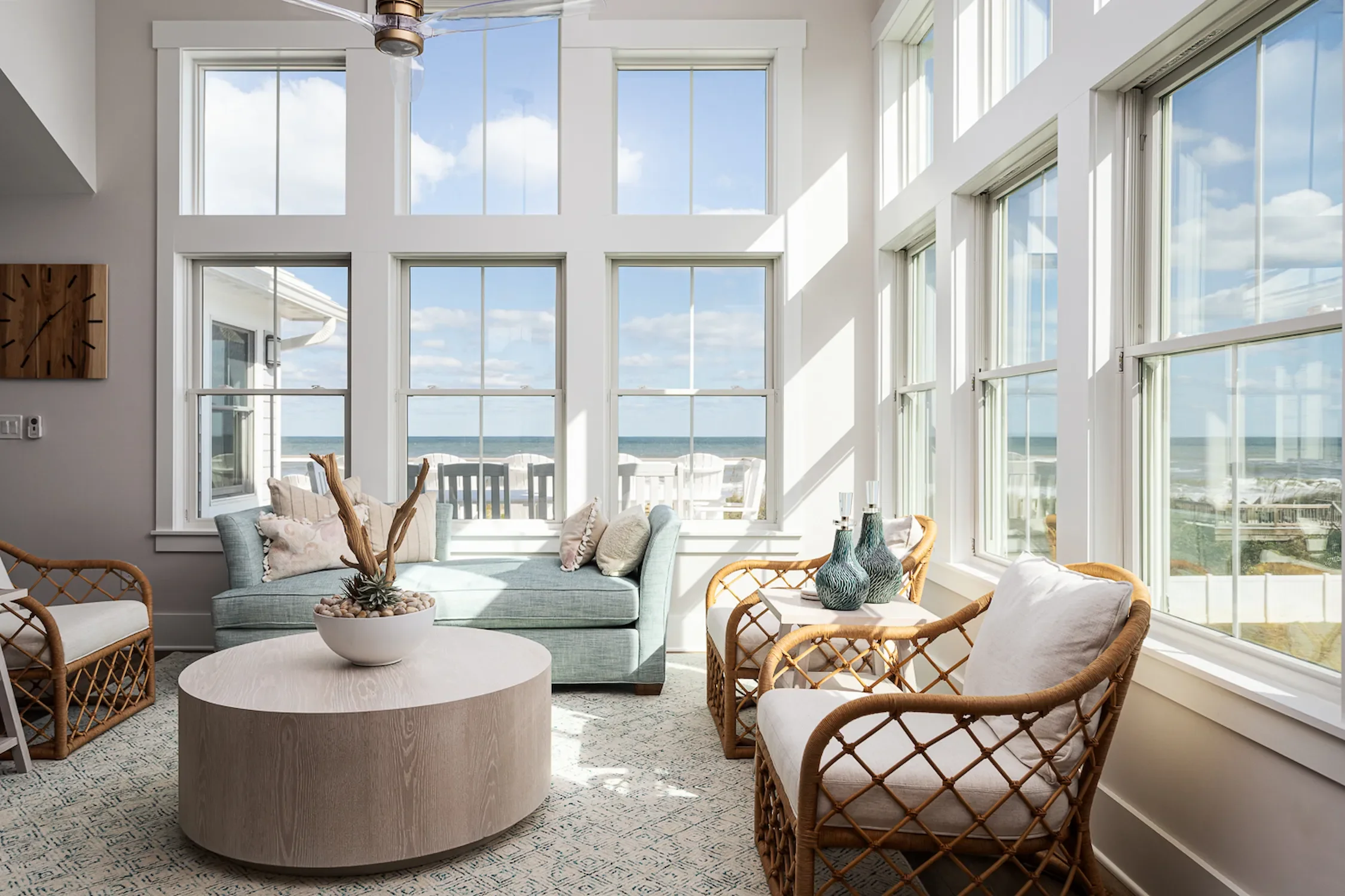 Sunlit coastal living room with large windows, wicker chairs, a sofa with pillows, a round coffee table with a decorative centerpiece, and ocean view.