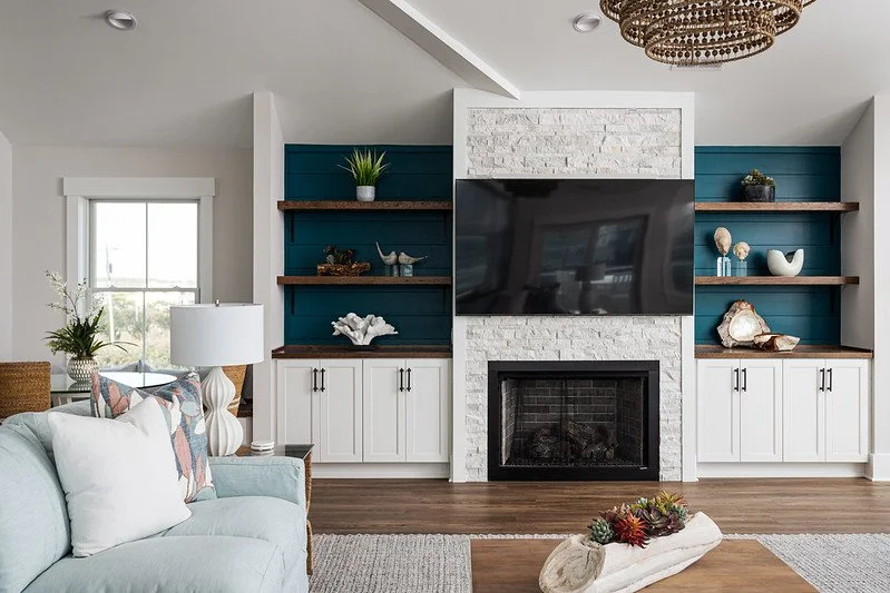 Living room with a fireplace, flat-screen TV above, blue built-in shelves with decorative items, white cabinetry, window on the left, and a light-colored sofa with pillows. A wooden coffee table with a plant arrangement is in the foreground.